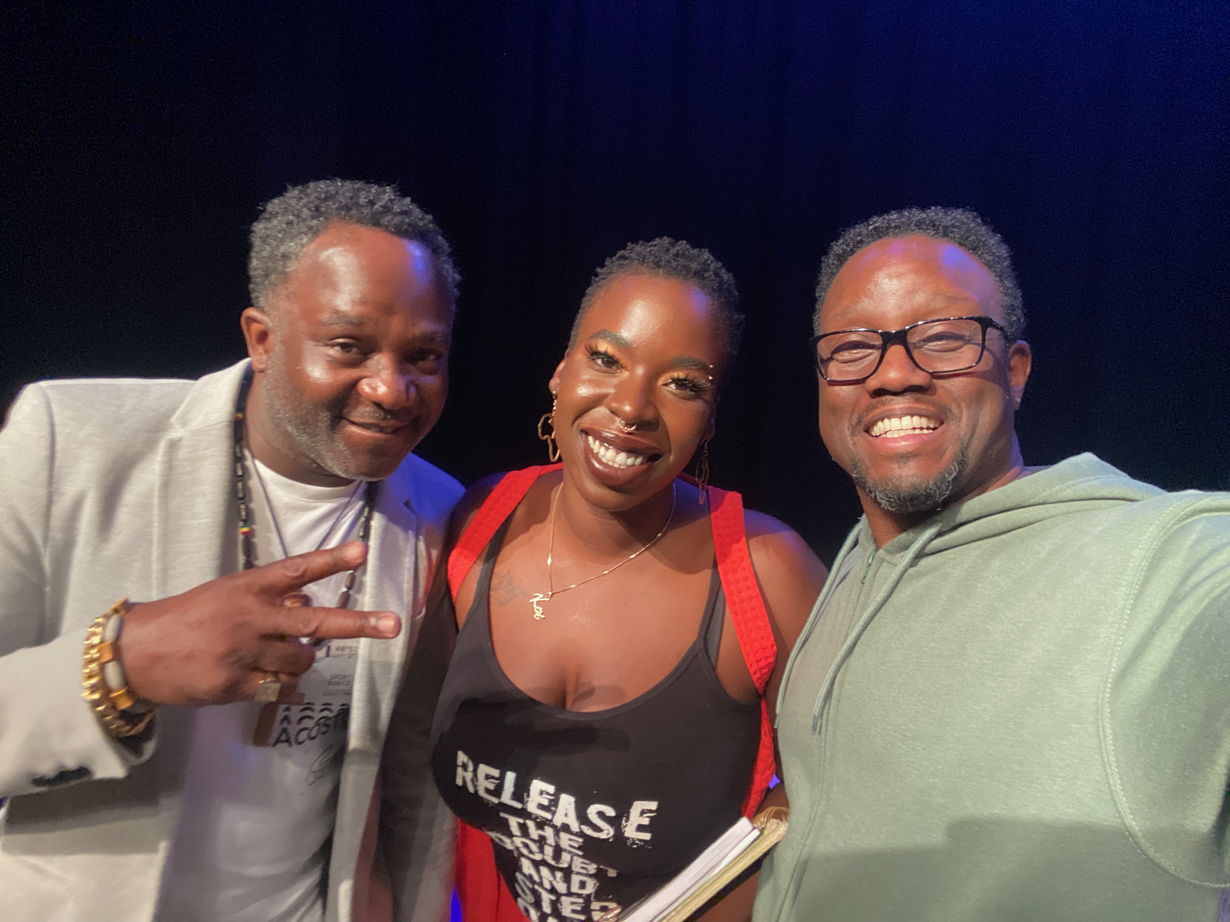 Three people smiling and taking a selfie together. The person on the left is a man with short, curly hair, wearing a light-colored blazer and making a peace sign. The person in the middle is a woman with short hair, wearing a black shirt with white t