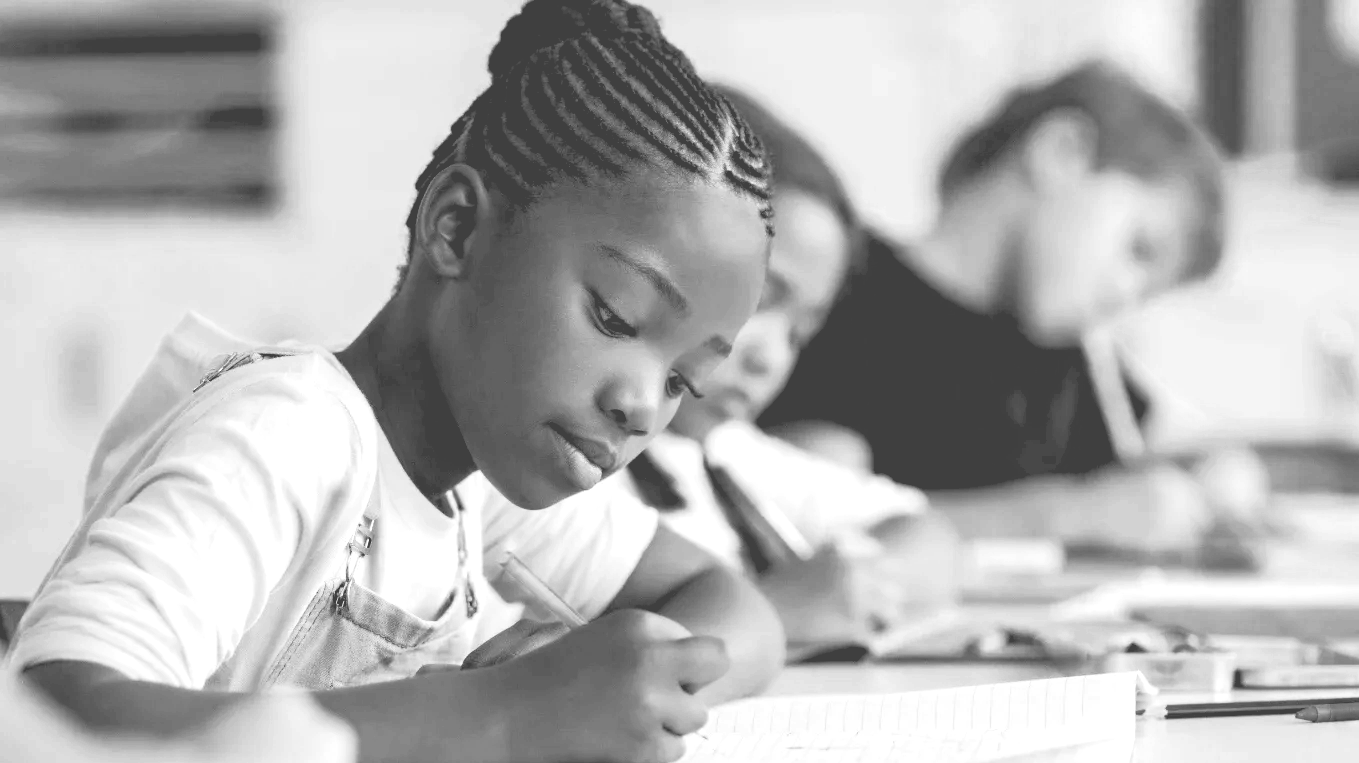 Young girl with braided hair writing in a notebook at a table, with other children in the background, in a classroom setting.