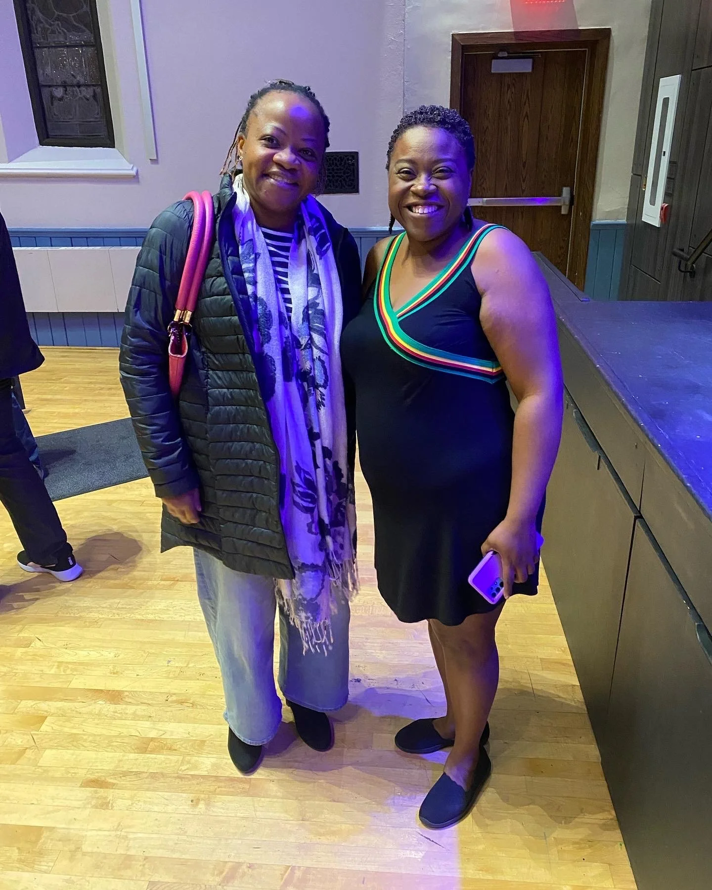 Two women standing together indoors, smiling at the camera. One woman is wearing a black dress with rainbow-colored straps, holding a smartphone, with a short natural hairstyle. The other woman is wearing a striped shirt, a black quilted jacket, a co