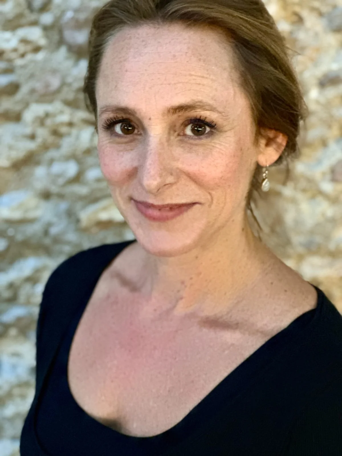 Portrait d'une femme souriante avec des cheveux bruns courts, portant un top noir, devant un mur en pierre beige.