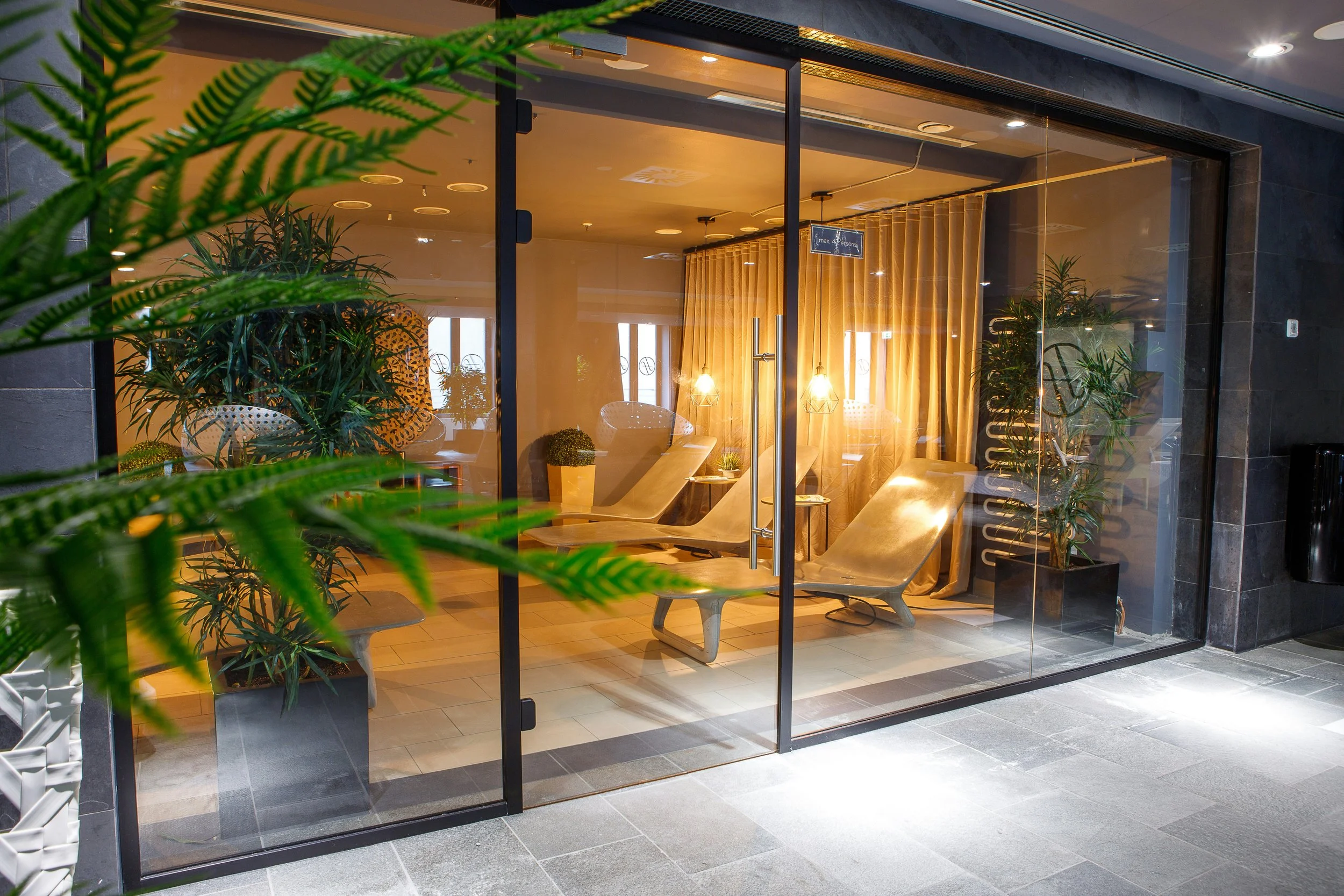 Intérieur d'un centre de bien-être ou spa avec des fauteuils en bois, des plantes vertes, des lumières douces et des rideaux beige derrière une grande baie vitrée.