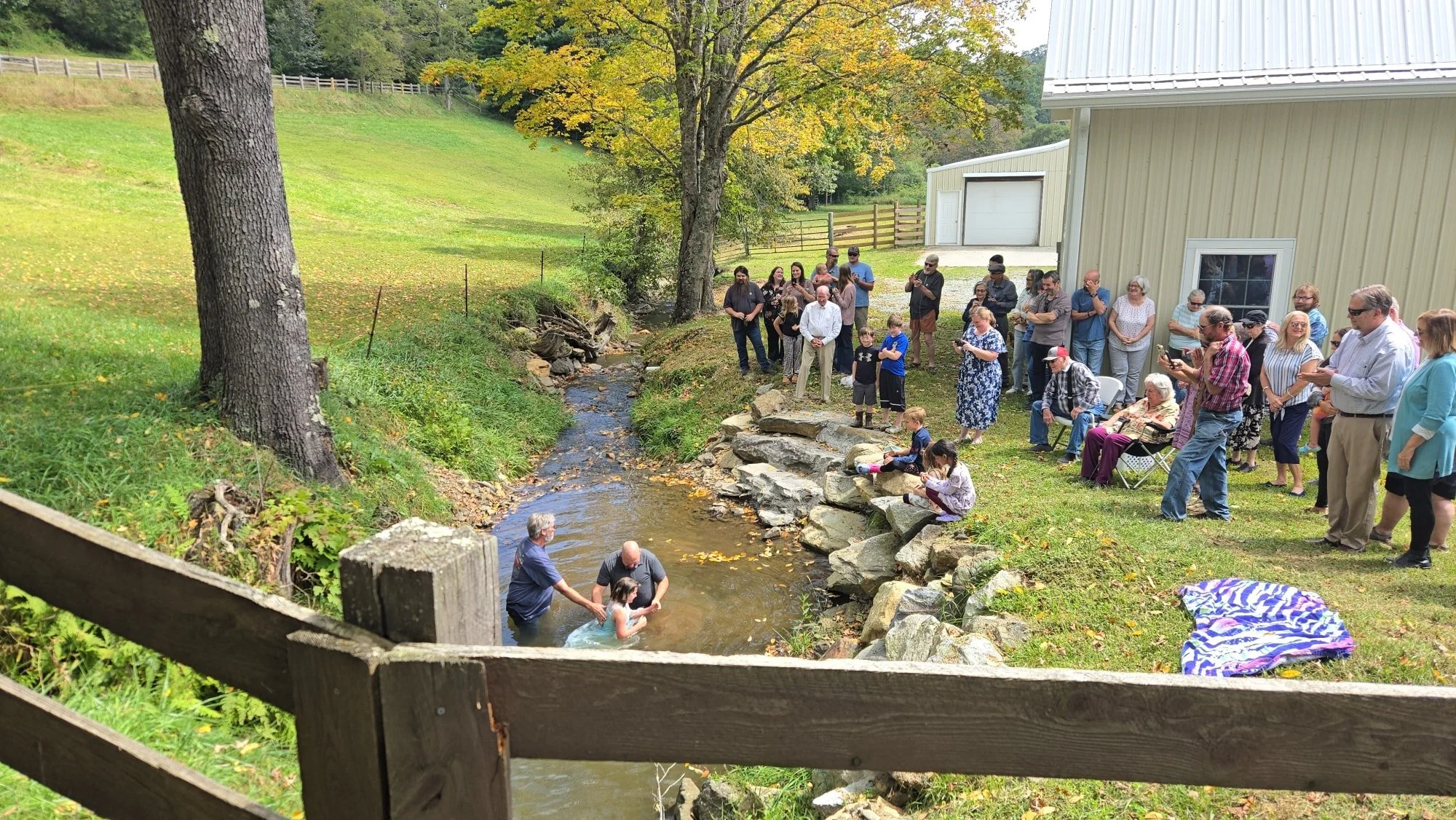 A group of people gathered outdoors along a small creek, some standing and some sitting on rocks, observing a baptism ceremony. The setting includes green grass, trees, and a large building with metal siding in the background.