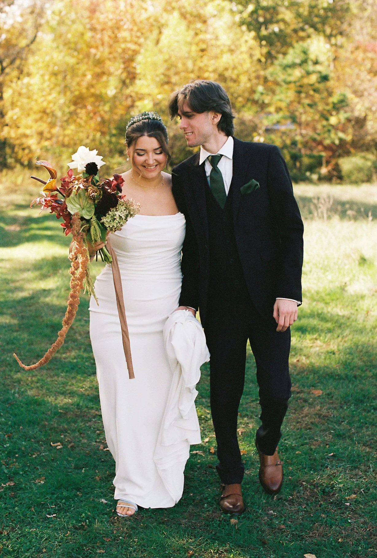 A bride in a white wedding dress holding a bouquet and a groom in a black suit walking outdoors on a grassy area with colorful autumn trees in the background.