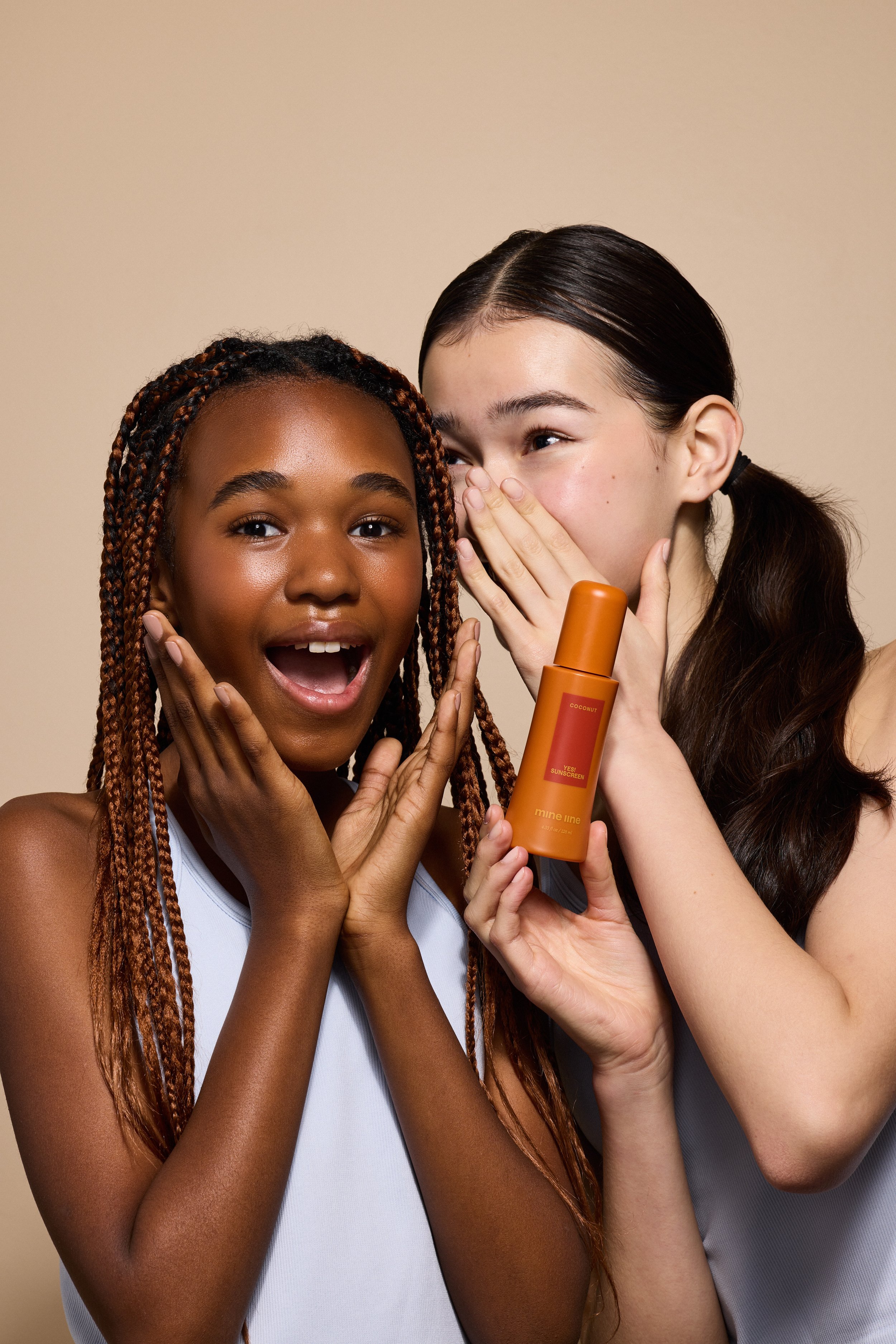 Two women enjoying a moment together, one holding a bottle of sunscreen, smiling and laughing.