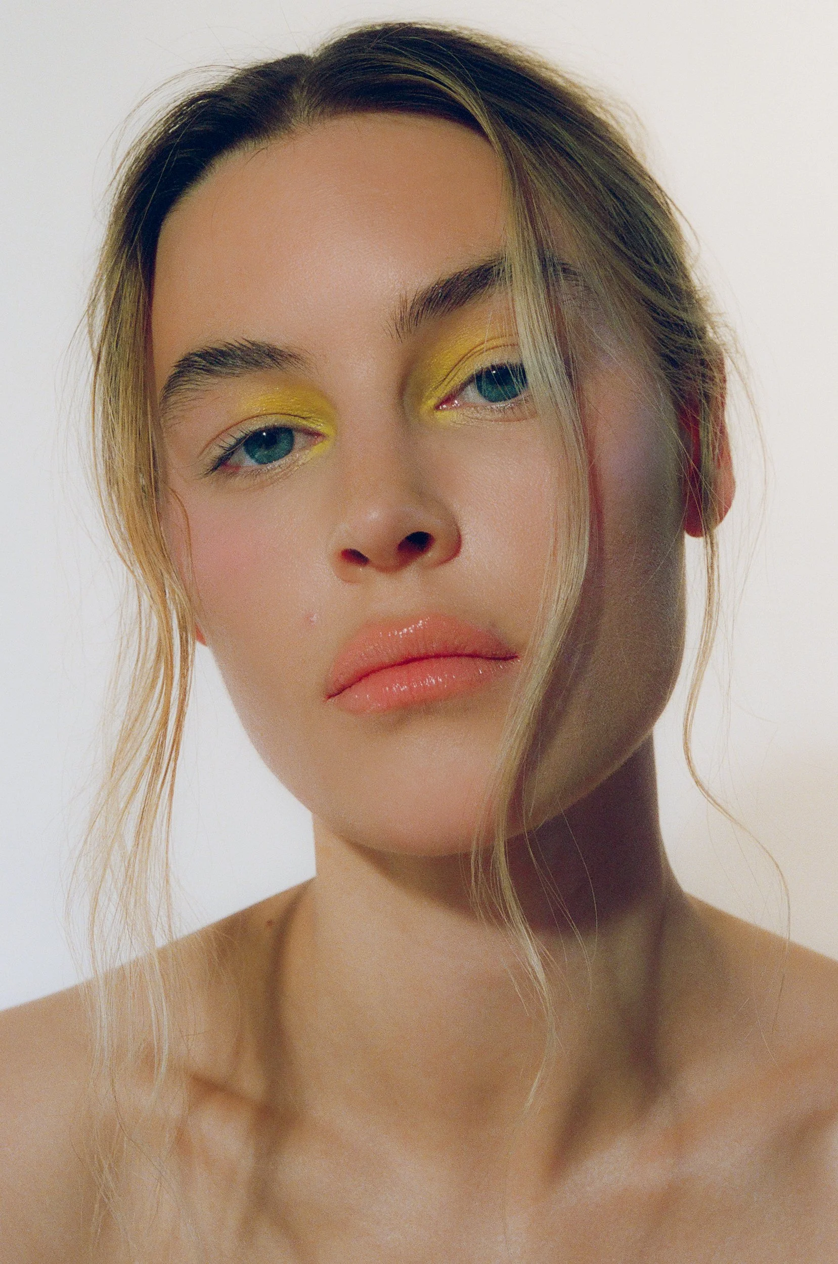Close-up portrait of a woman with yellow eyeshadow, natural makeup, and loose hair, looking at the camera with a calm expression.