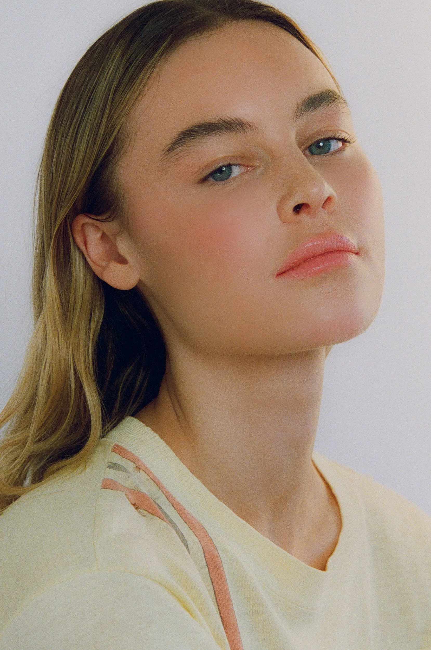 Close-up of a woman's face with light makeup, blue eyes, and wavy blonde hair, wearing a light yellow top with pink and gray accents.