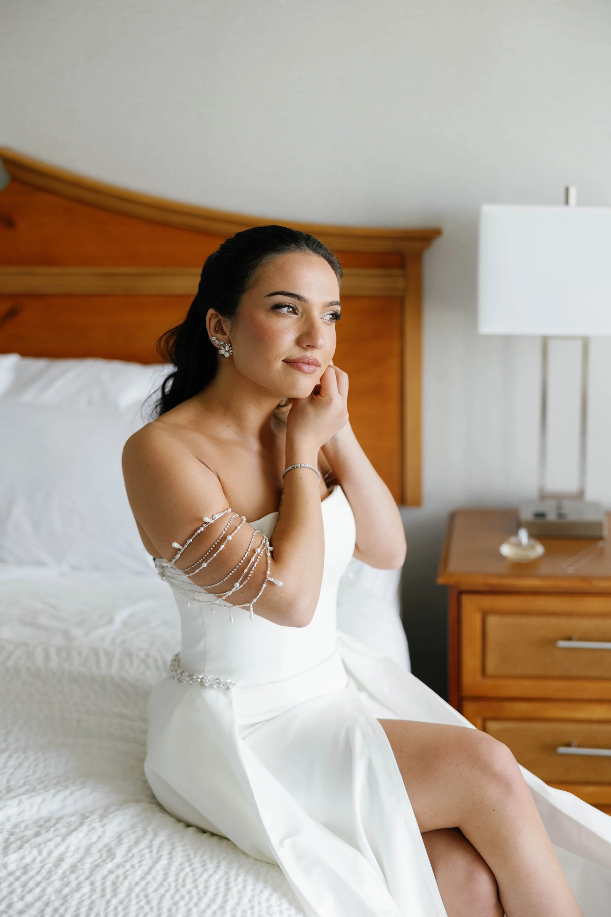 Bride sitting on a bed, adjusting her earring, dressed in a white wedding gown with pearl jewelry, in a hotel room.