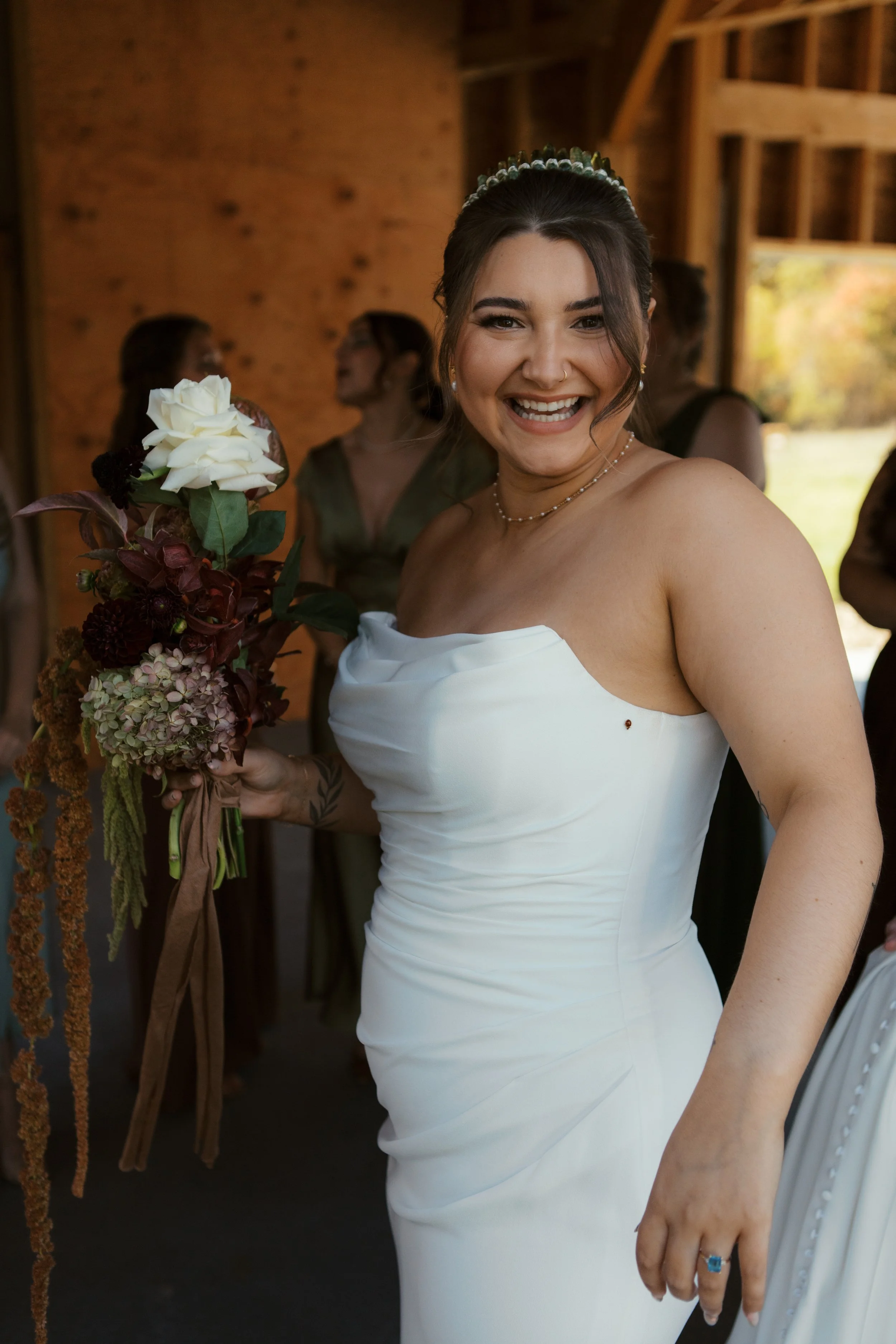 A bride in a strapless white wedding dress holding a bouquet of flowers, smiling happily, with bridal party members in the background inside a rustic venue.