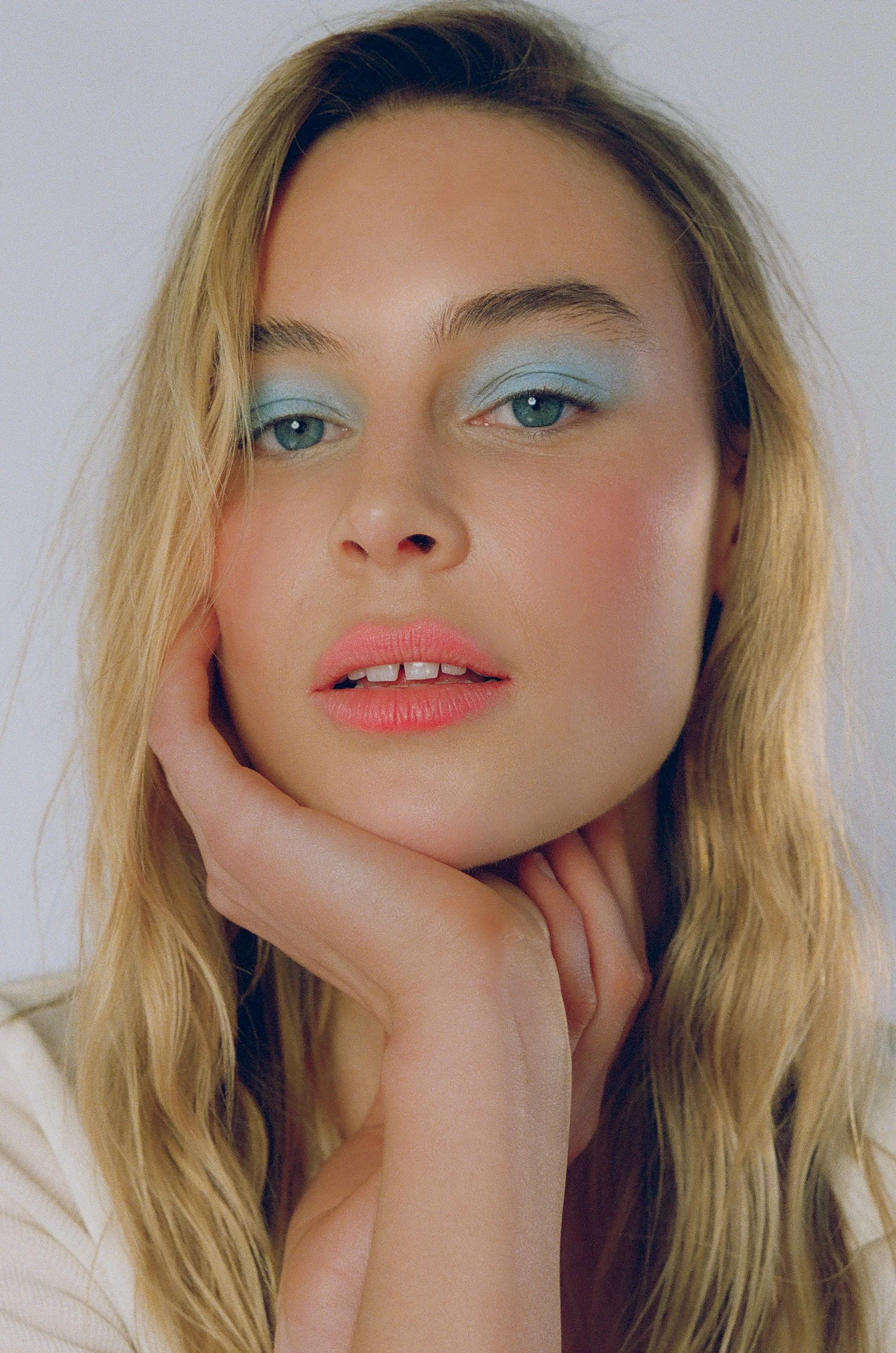 A close-up portrait of a woman with blue eyes and blonde wavy hair. She has pink lipstick, blue eyeshadow, and is resting her face on her hand.