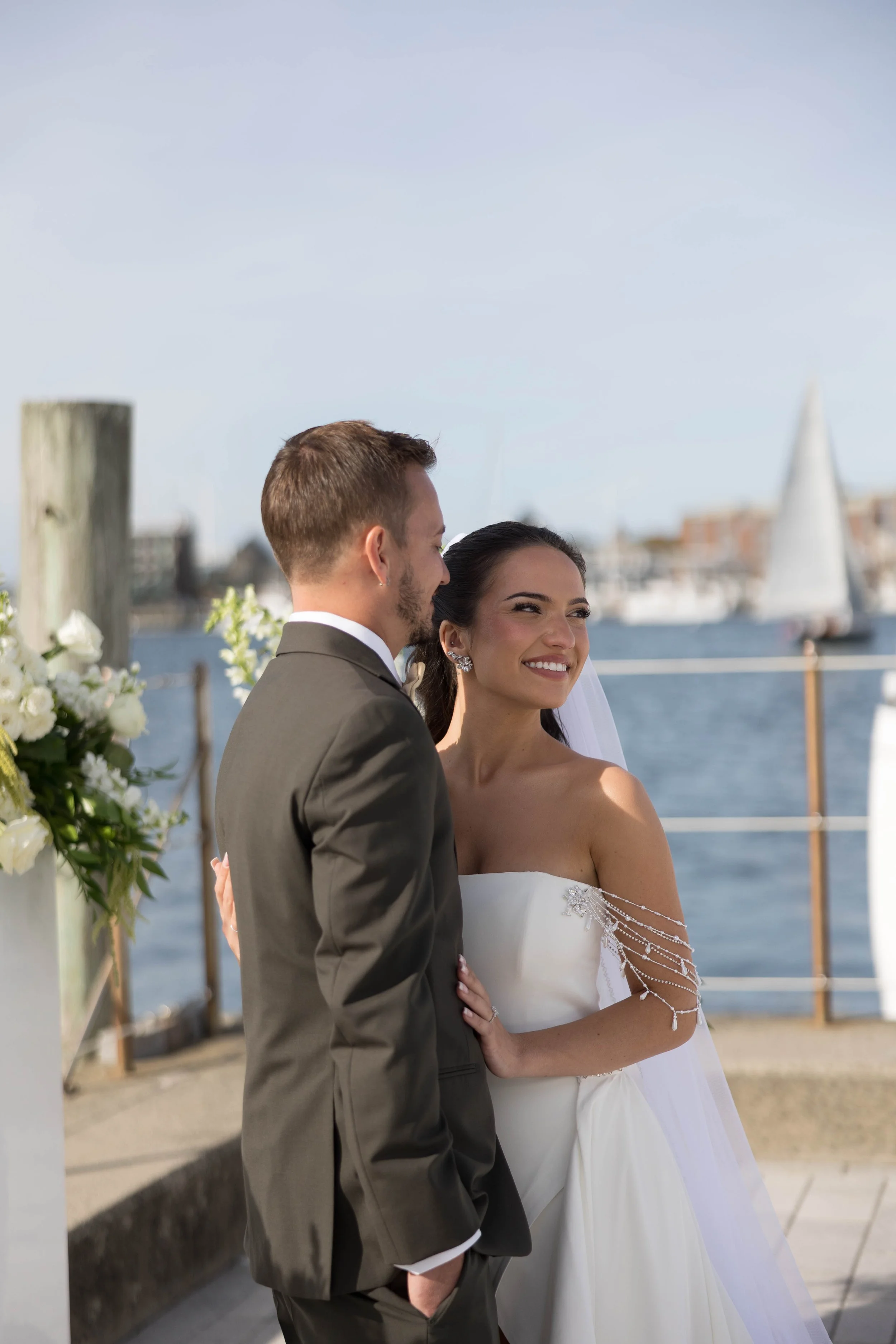 Bride and groom standing together outdoors near water, smiling with sailboats and buildings in the background.