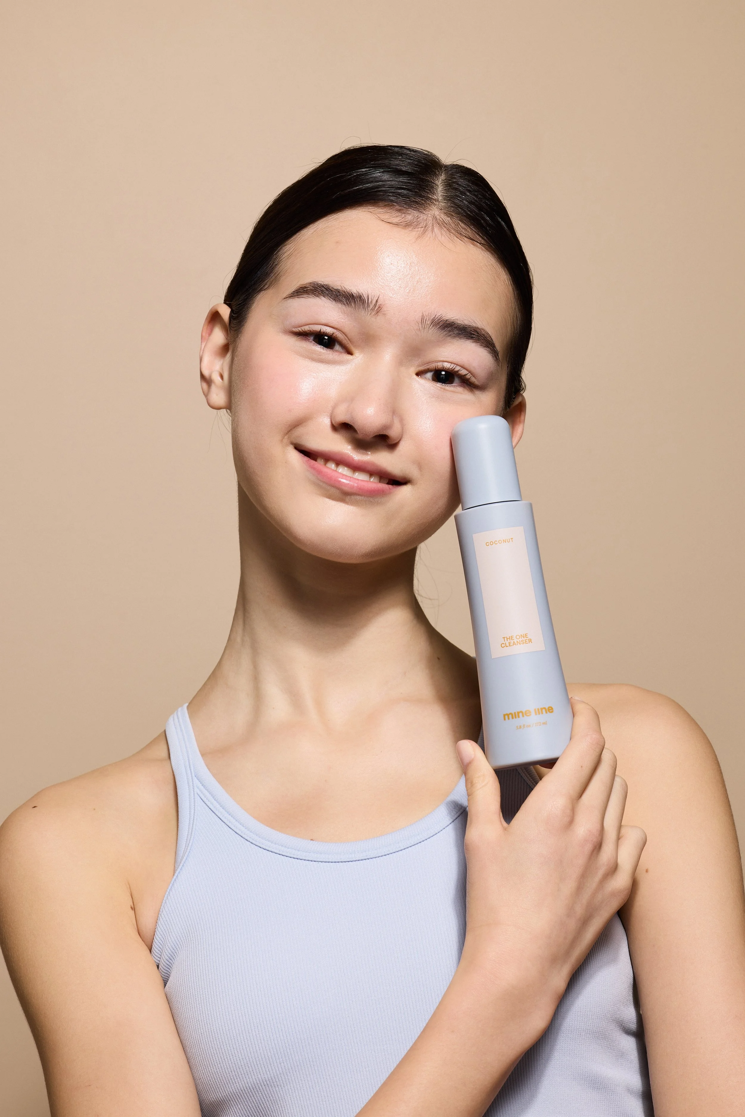 A young woman with dark hair and light skin holding a bottle of skincare product labeled 'mine line' and 'Coconut,' smiling at the camera against a beige background.