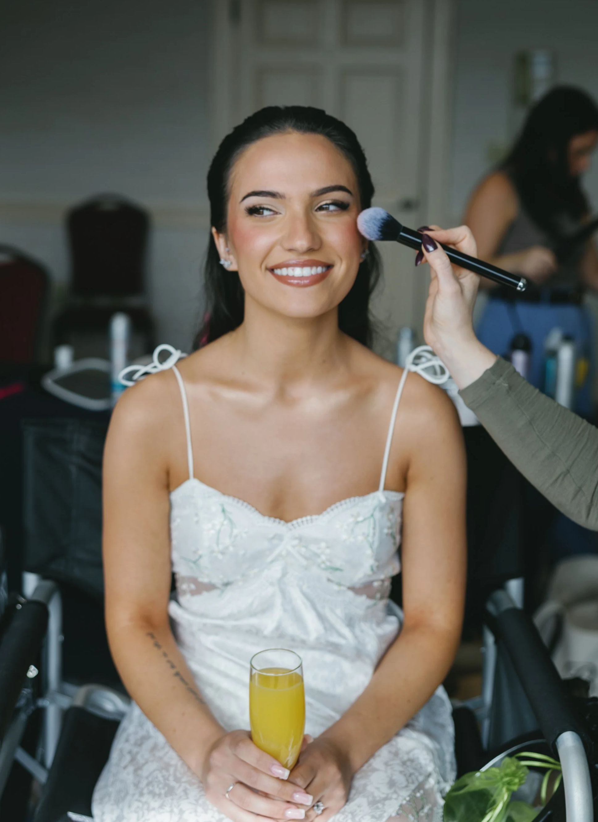 Bride in a white dress smiling as makeup artist applies makeup, holding a glass of orange juice.