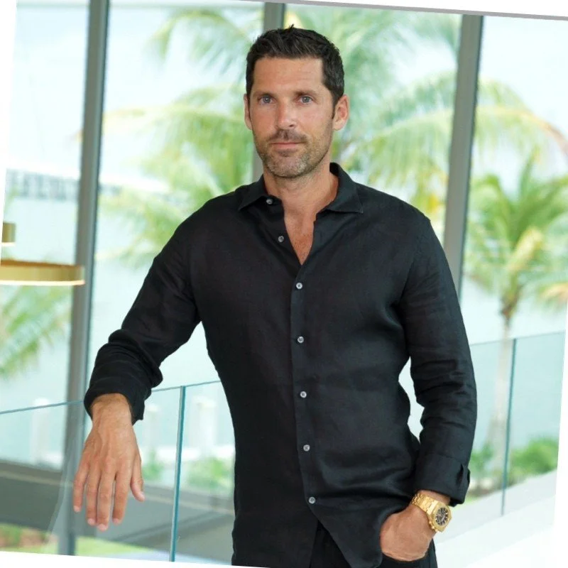 A man with short dark hair, beard, and light skin wearing a black button-up shirt and a gold watch, standing near large windows with green foliage outside, in a modern indoor setting.