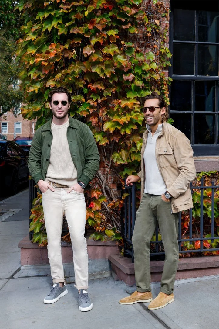 Two men standing on a sidewalk in front of a ivy-covered brick wall and a window, both wearing sunglasses and casual jackets.