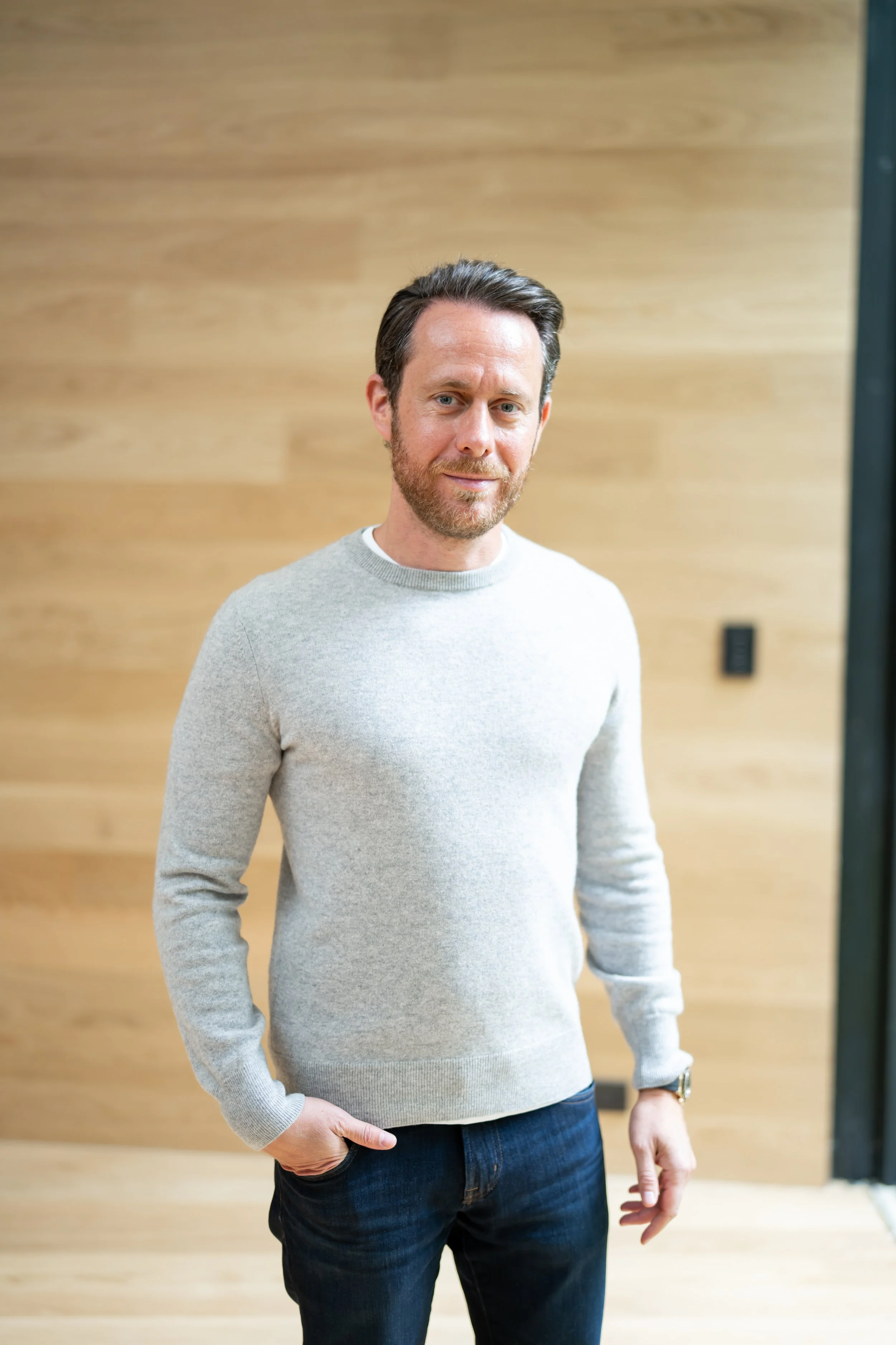 A man standing indoors against a wooden wall, wearing a light gray sweater and dark jeans, with one hand in his pocket.