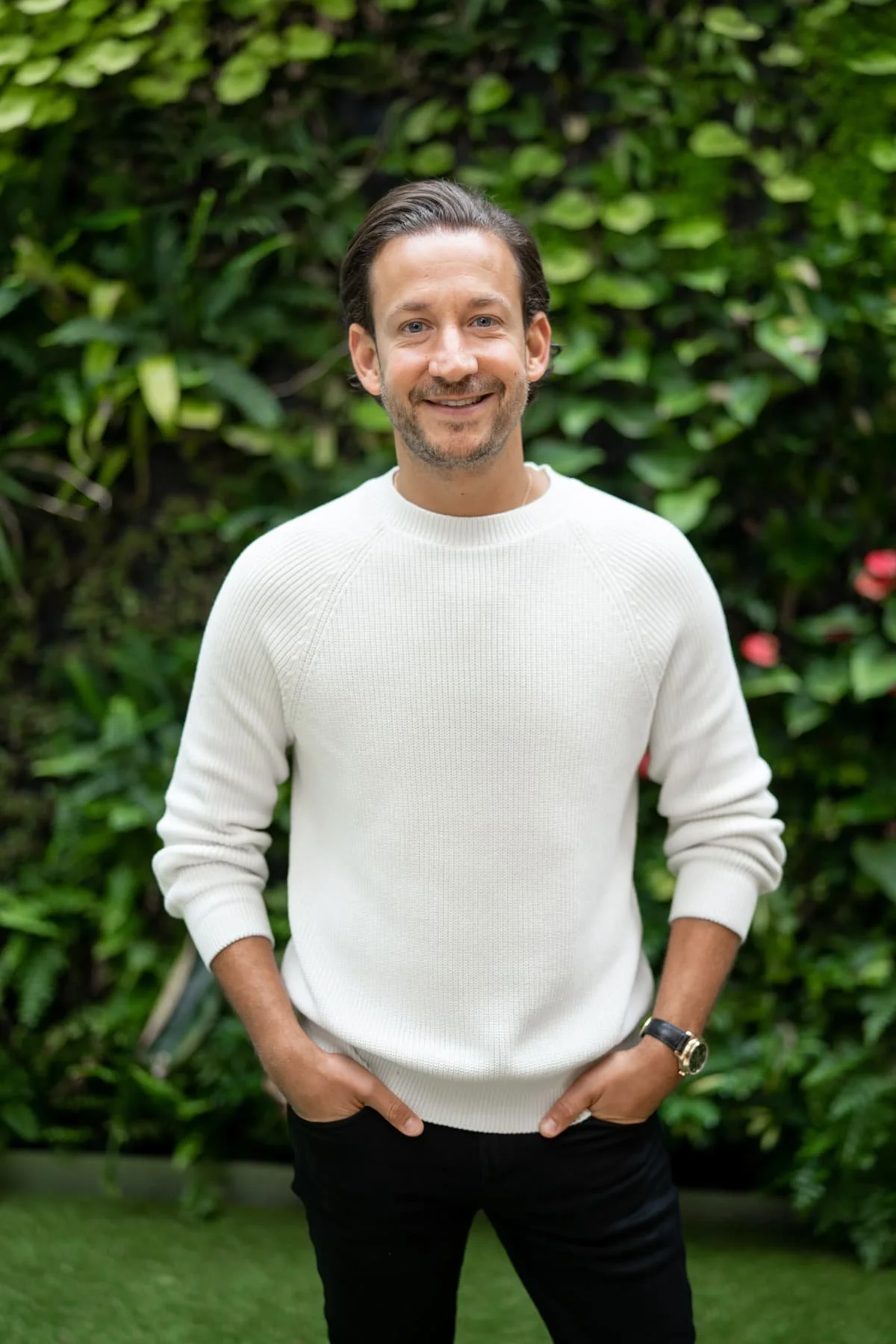 A smiling man in a white sweater and black pants stands outdoors with his hands in his pockets, against a background of green plants and foliage.