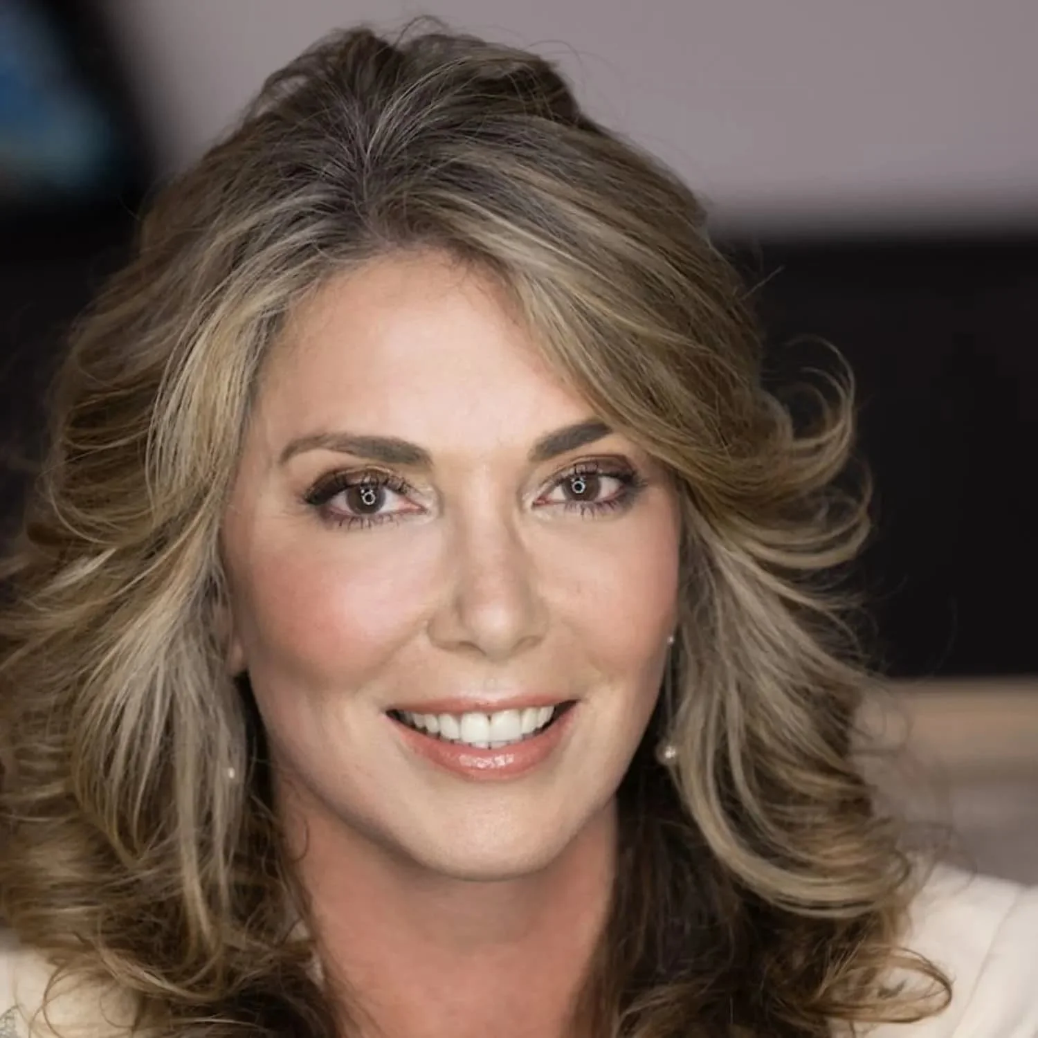 Close-up of a smiling woman with wavy blonde hair and makeup, wearing pearl earrings.