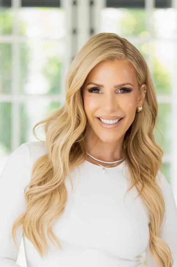 A woman with long blonde hair smiling, wearing a white top and jewelry, standing in front of a window with a green outdoor background.