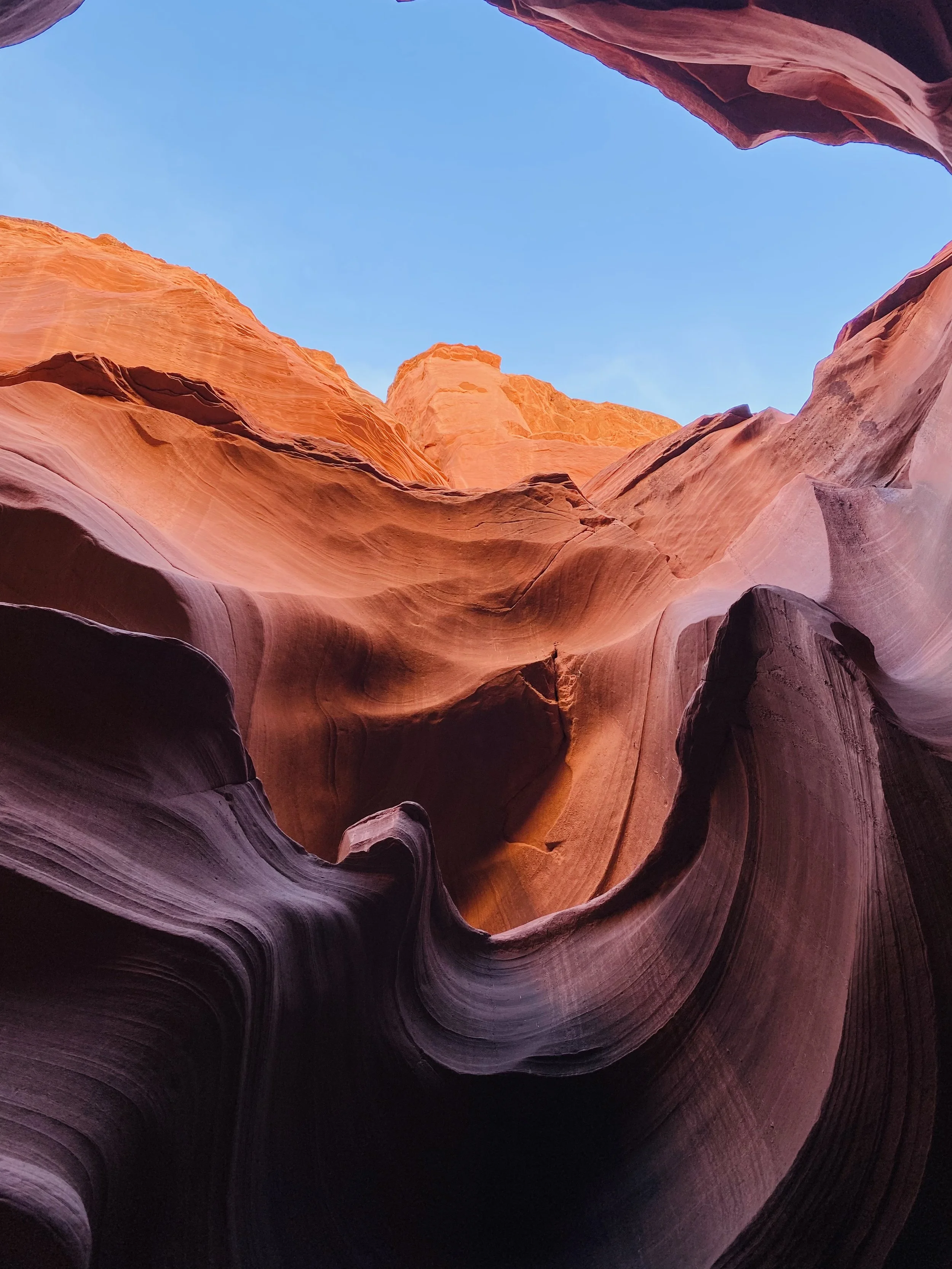 View looking up into a slot canyon with smooth, colorful sandstone walls and a clear blue sky overhead.
