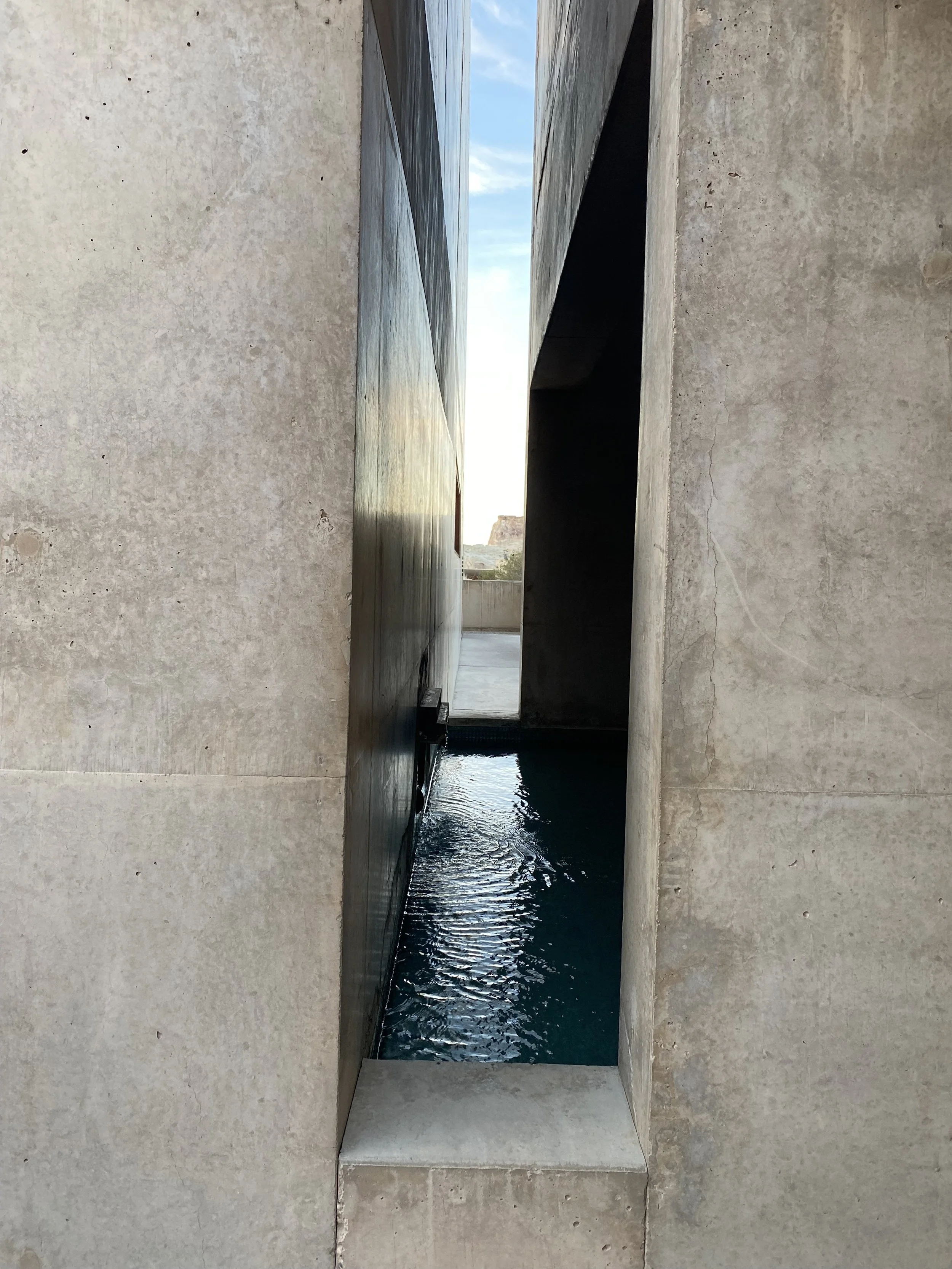 Modern architectural structure with concrete walls and narrow opening revealing a water feature and outdoor sky.