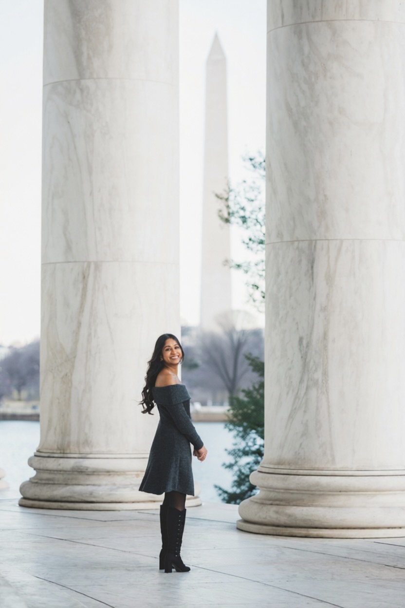 A woman in a black off-the-shoulder dress and tall black boots standing between two large marble columns with the Washington Monument in the background.
