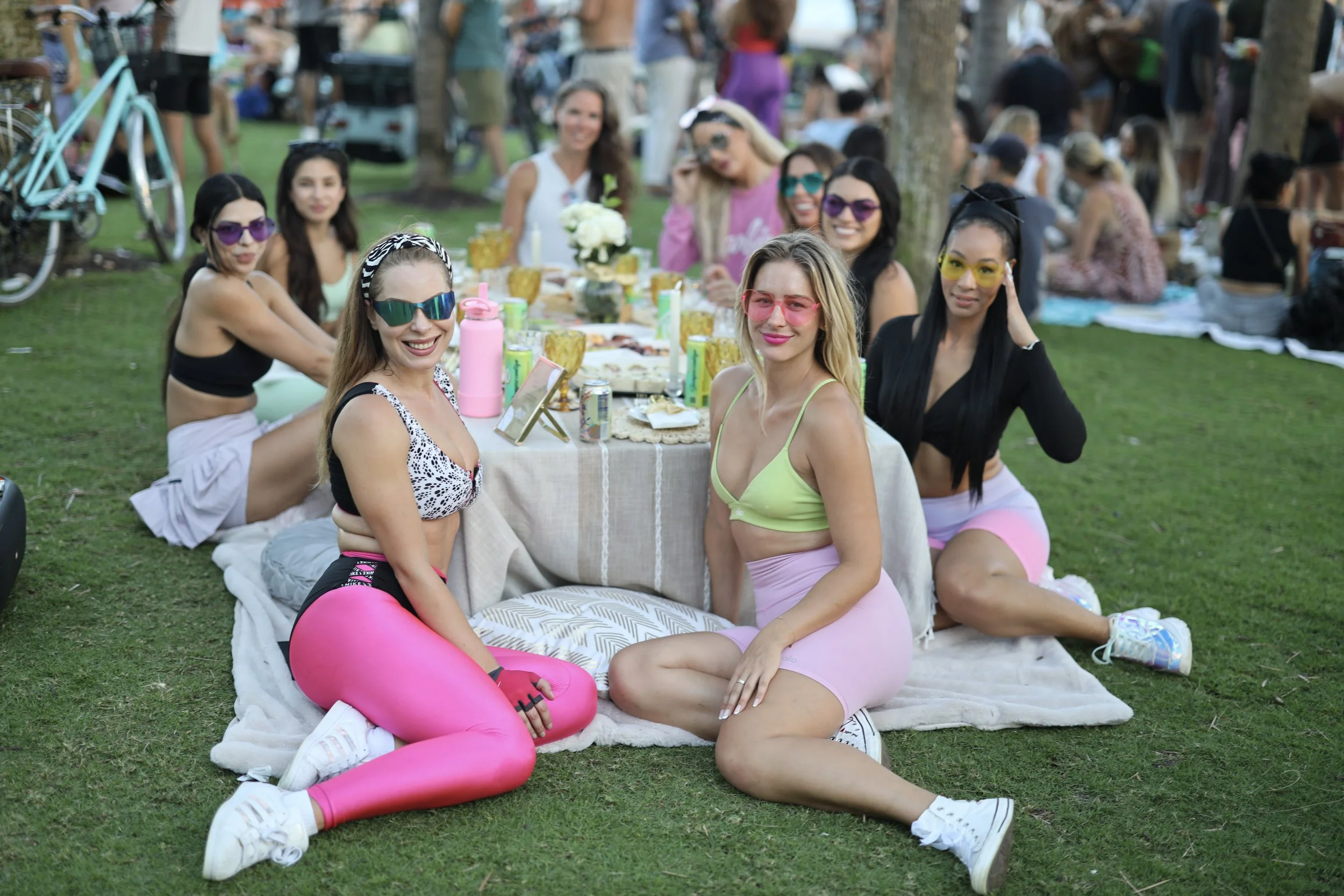 Group of women having a picnic outdoors at a park, sitting on a blanket with a table set with food and drinks, all wearing colorful casual and athletic clothing with sunglasses.