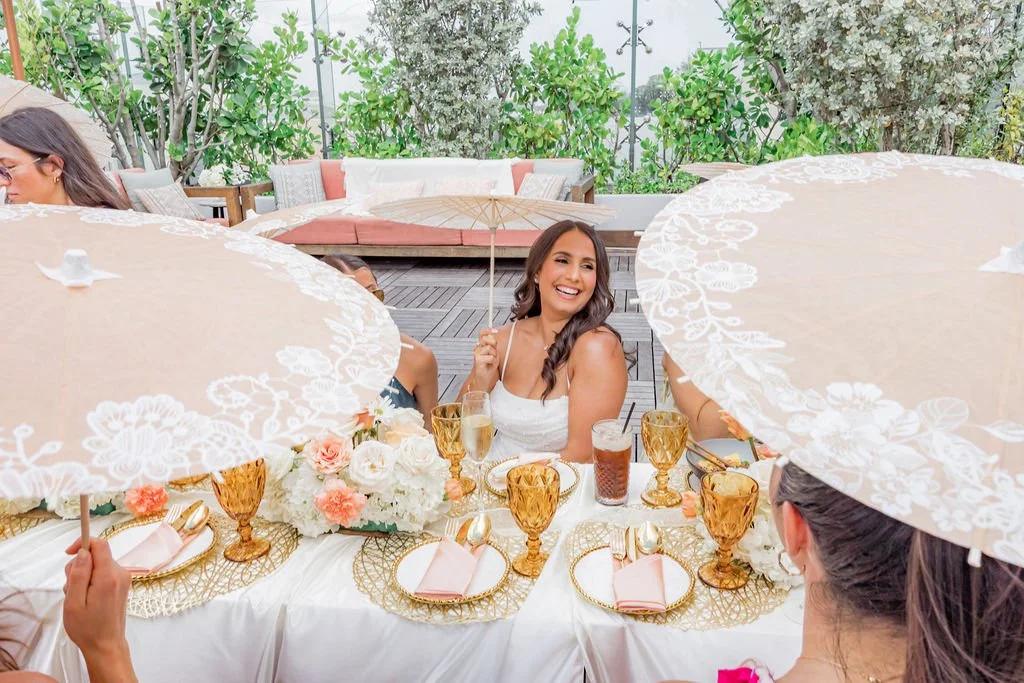A woman sitting at a decorated outdoor table during a celebration, smiling and holding a paper parasol, with other women partially visible holding lace parasols around her.