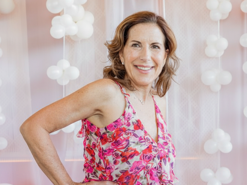 A woman with shoulder-length brown hair smiling, wearing a pink floral sleeveless top, standing in front of a backdrop with white spherical decorations.