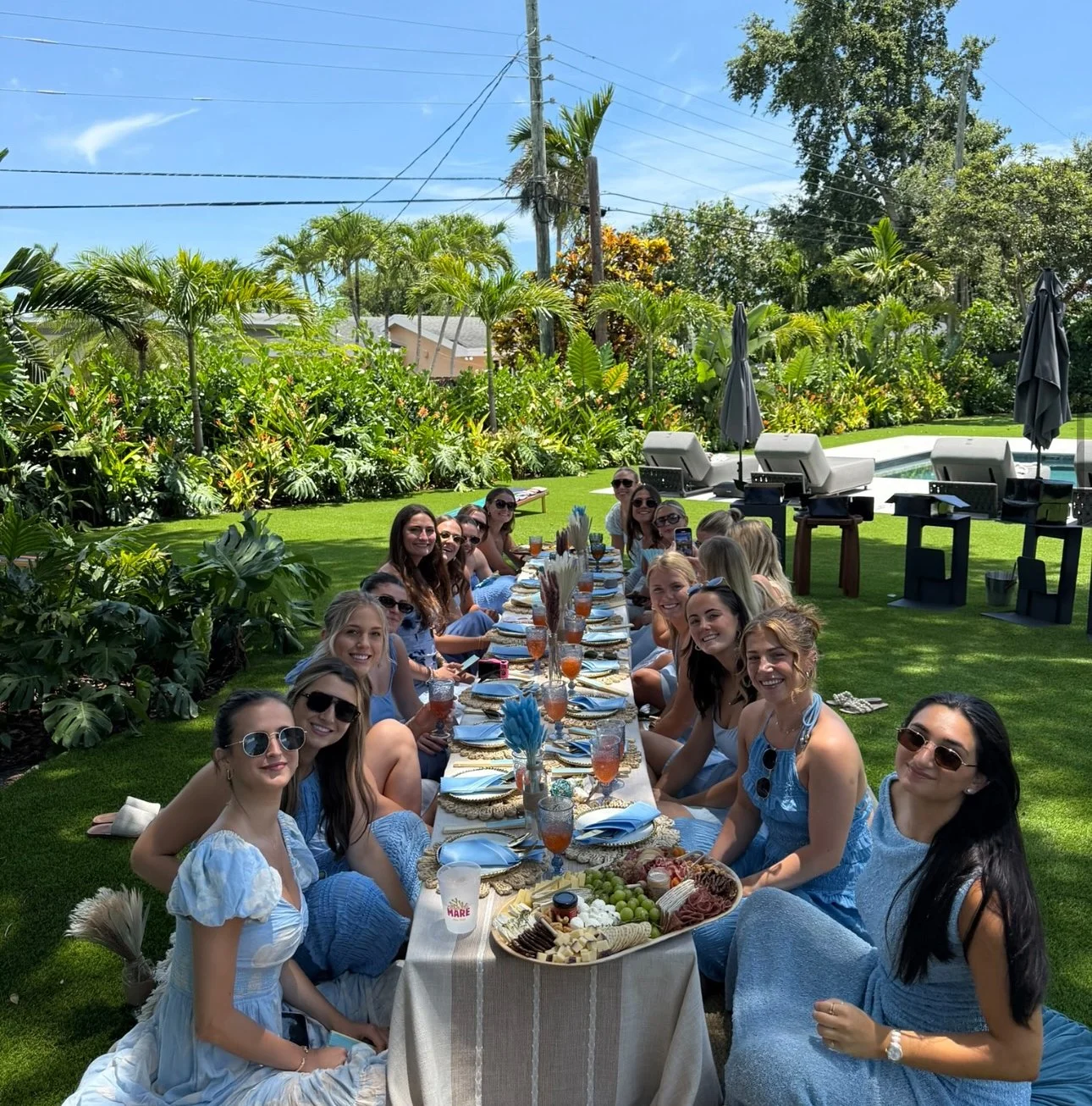 A group of women gathered around a long outdoor table on a sunny day, enjoying a meal in a lush, green backyard with a swimming pool, lounge chairs, and tropical plants.