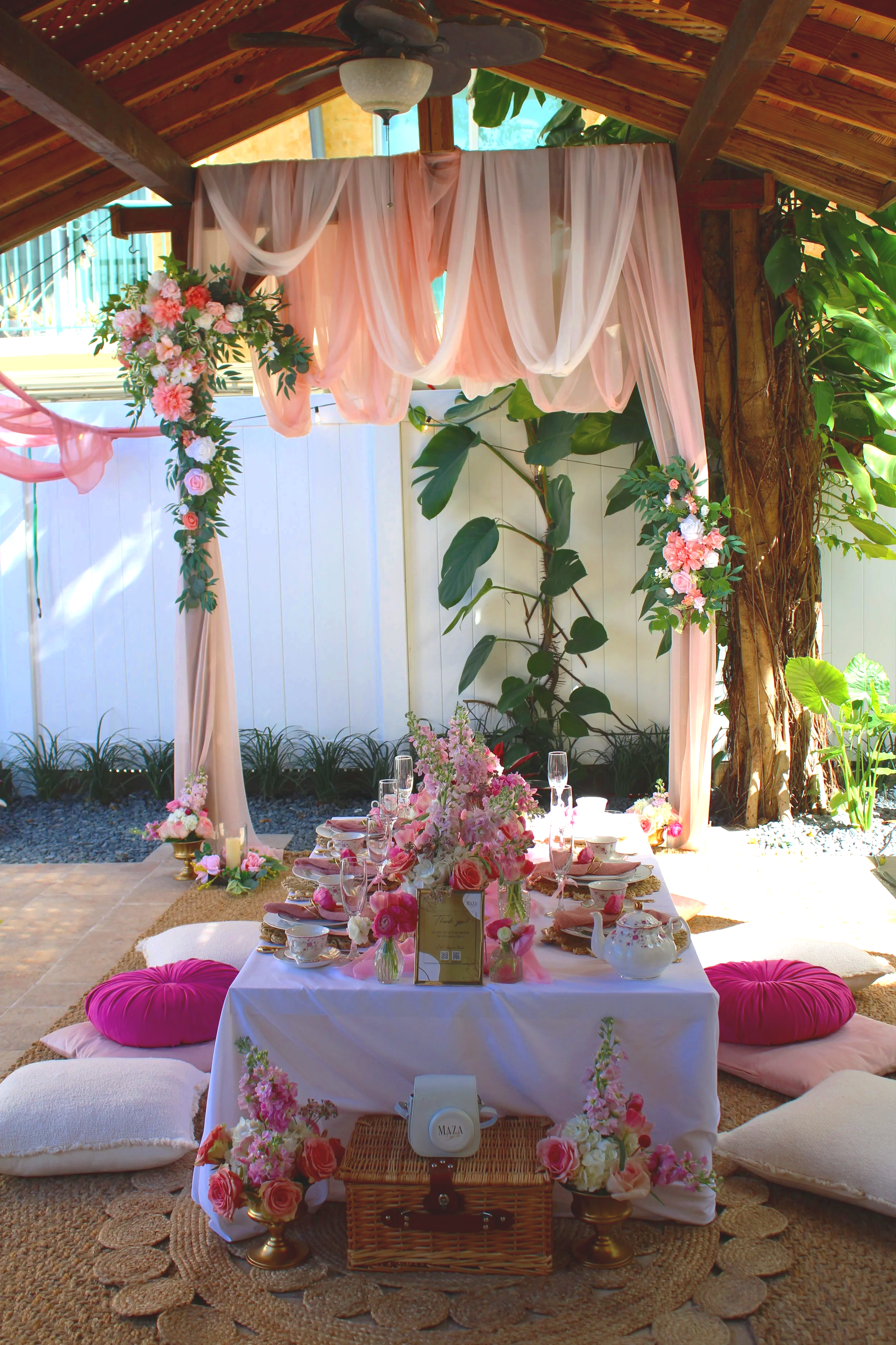 Decorated outdoor seating area for an event with a table set with pink and white flowers, pink cushions, and a pink fabric canopy, surrounded by lush greenery.