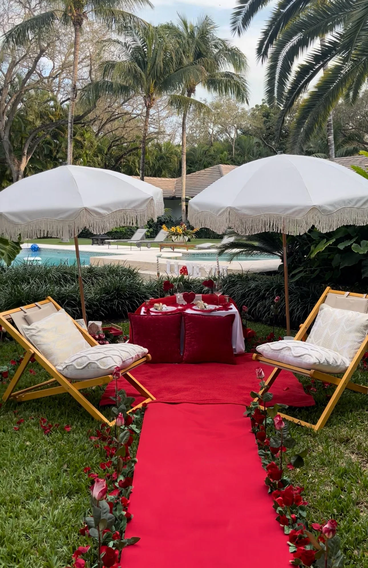 A romantic outdoor setting with a red carpet walkway lined with pink and red roses, leading to a table for two with heart-shaped décor. The scene is shaded by two white umbrellas and surrounded by lounge chairs, overlooking a pool and lush tropical t
