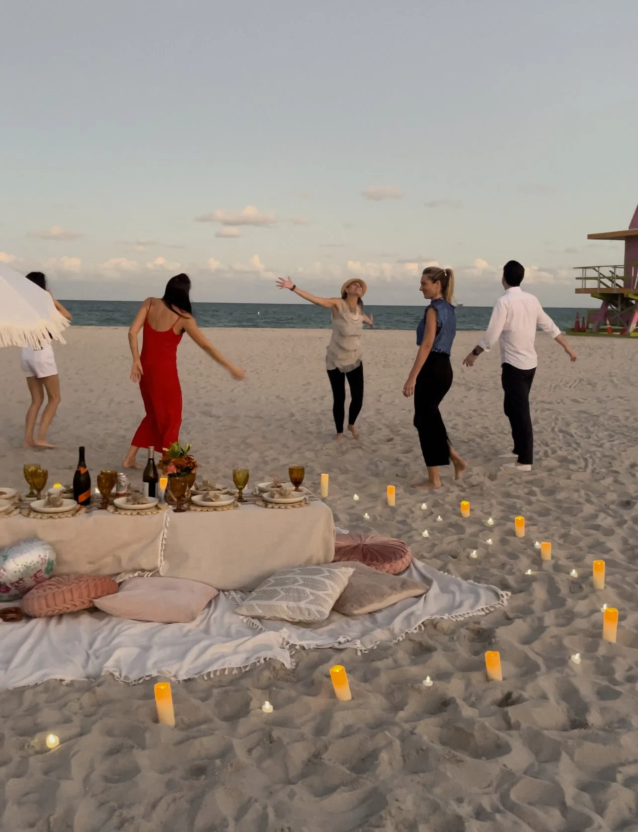 Group of people dancing on the beach during sunset, with a picnic setup including candles, pillows, wine bottles, and glasses.