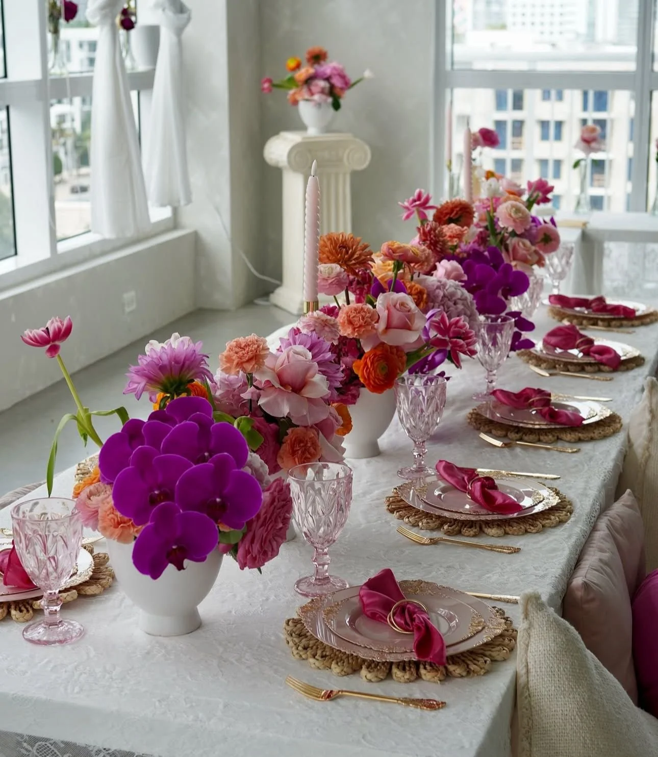 A decorated dining table with pink, purple, and orange flowers in white vases, pink candles, gold flatware, pink glassware, and pink napkins on plates. The table is set for a celebration in a bright room with large windows.