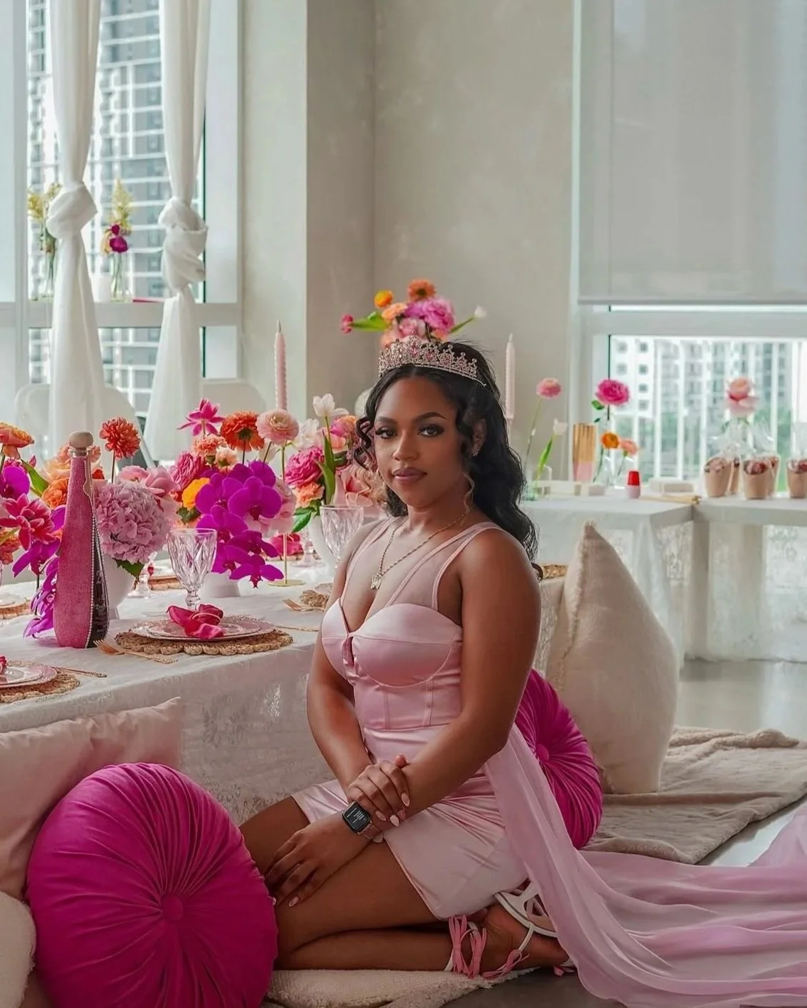 A young woman wearing a pink dress and a tiara sits on a cushion in a decorated room with pink and purple flowers, featuring a table set for a celebration with glassware and plates.