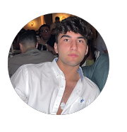 Young man with dark hair and eyebrows, wearing a white shirt, sitting indoors at a social event.