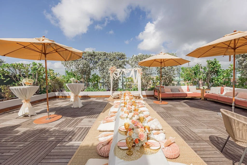 Setting for a rooftop wedding with a long table decorated with flowers and candles, surrounded by umbrellas and lounge seating, under a blue sky with clouds.