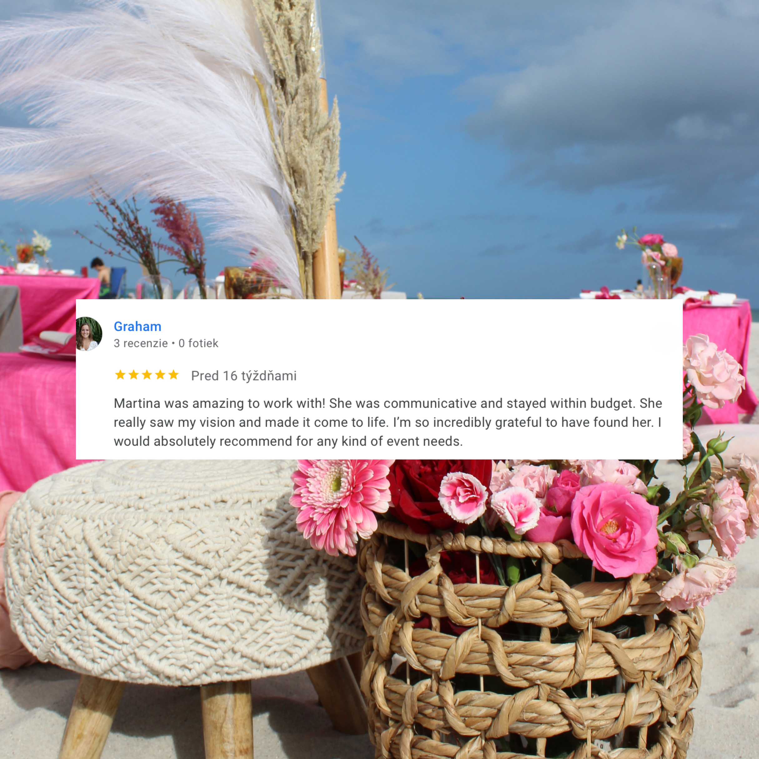 Beachside event setup with pink tablecloths, floral arrangements, a woven basket filled with pink and red flowers, a woven round ottoman, a basket with pampas grass, and an umbrella, under a partly cloudy sky.
