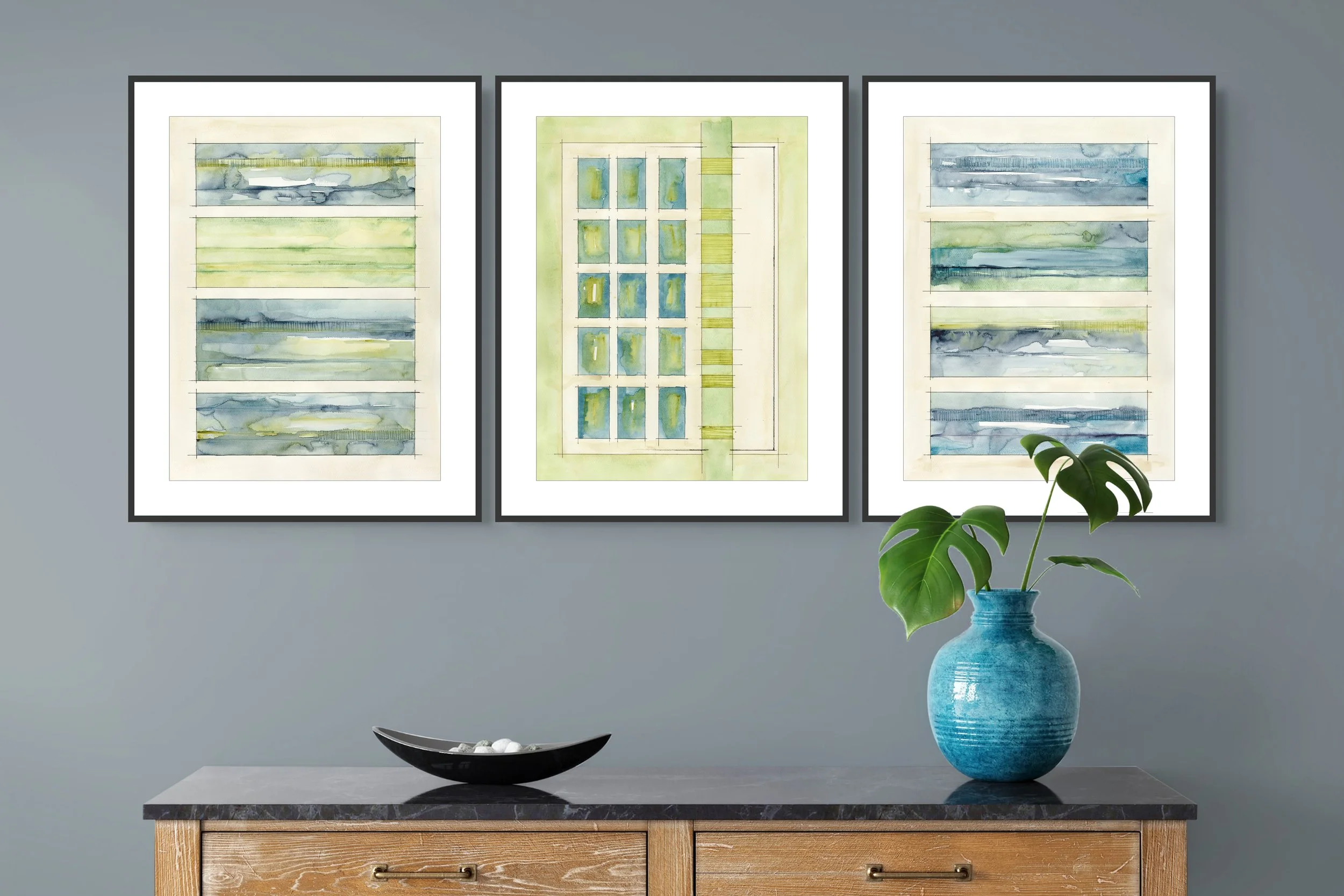 A wooden sideboard with a black marble top, a black decorative bowl, and a blue ceramic vase with green monstera leaves, positioned in front of a gray wall decorated with three abstract watercolor art pieces in black frames.