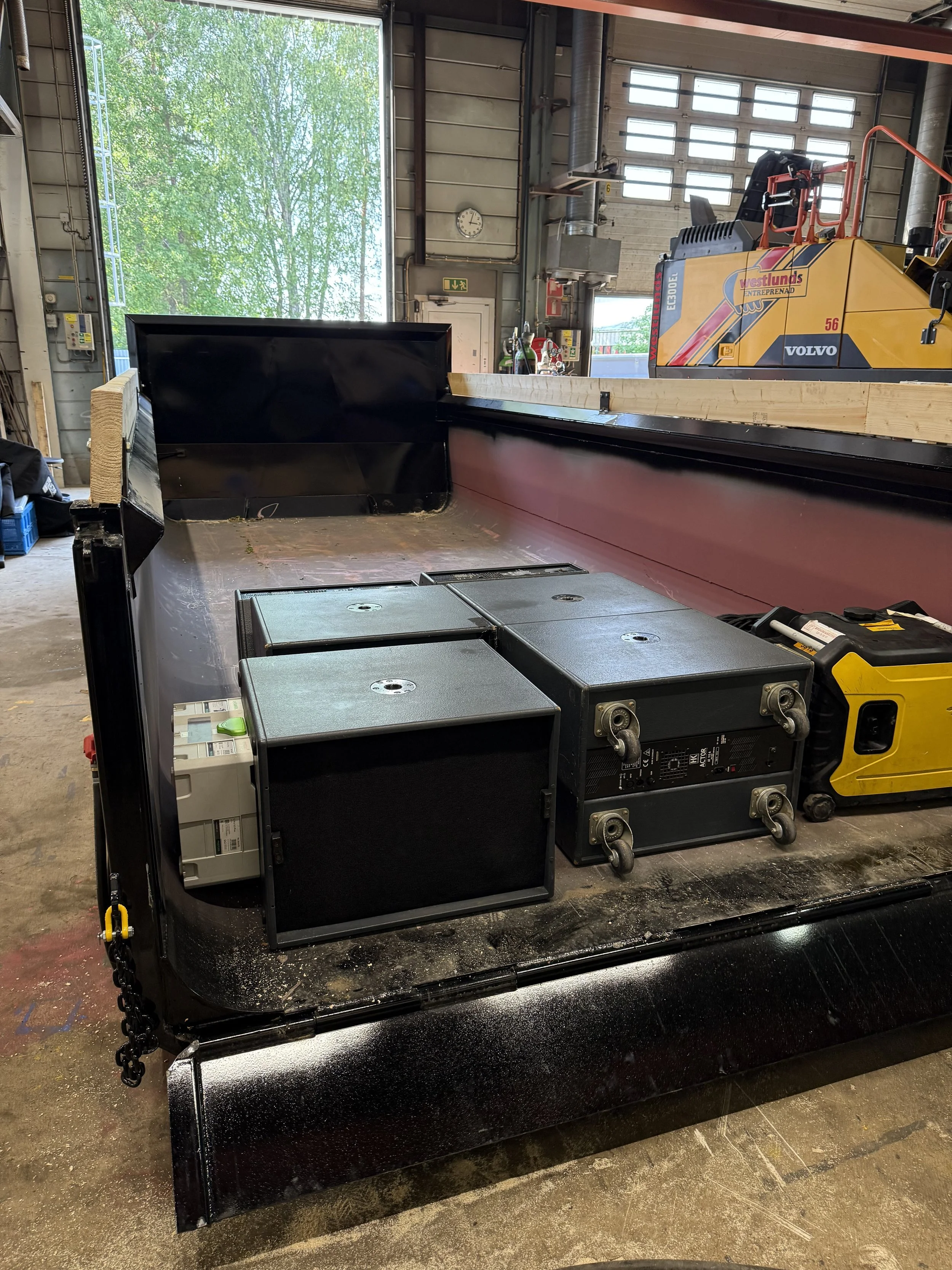 Inomhusmiljö med arbetsutrustning, inklusive svarta lådor och elektronisk utrustning, samt en bakgrund med en stor öppen dörr och ett ur som visar tiden.