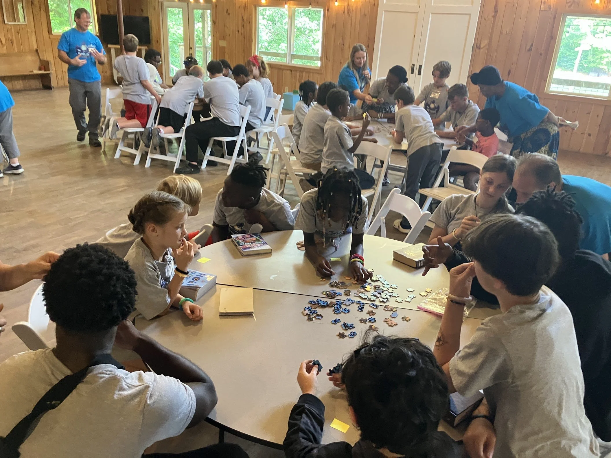 Group of children and adults playing puzzles and engaging in activities at tables inside a wooden cabin with large windows.