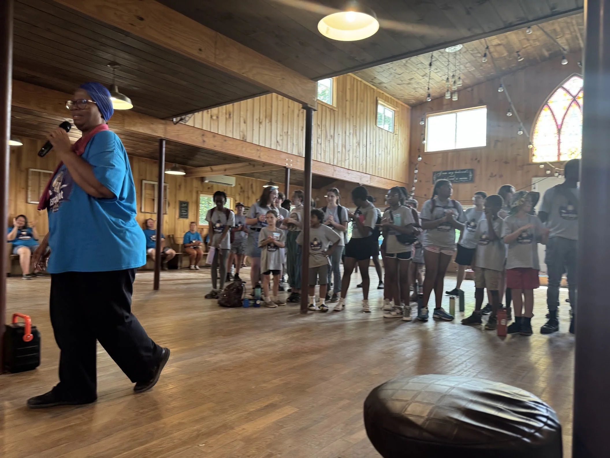 A woman in a blue shirt and headscarf holding a microphone speaking to a group of children and teenagers inside a wooden hall with sunlight coming through stained glass windows.