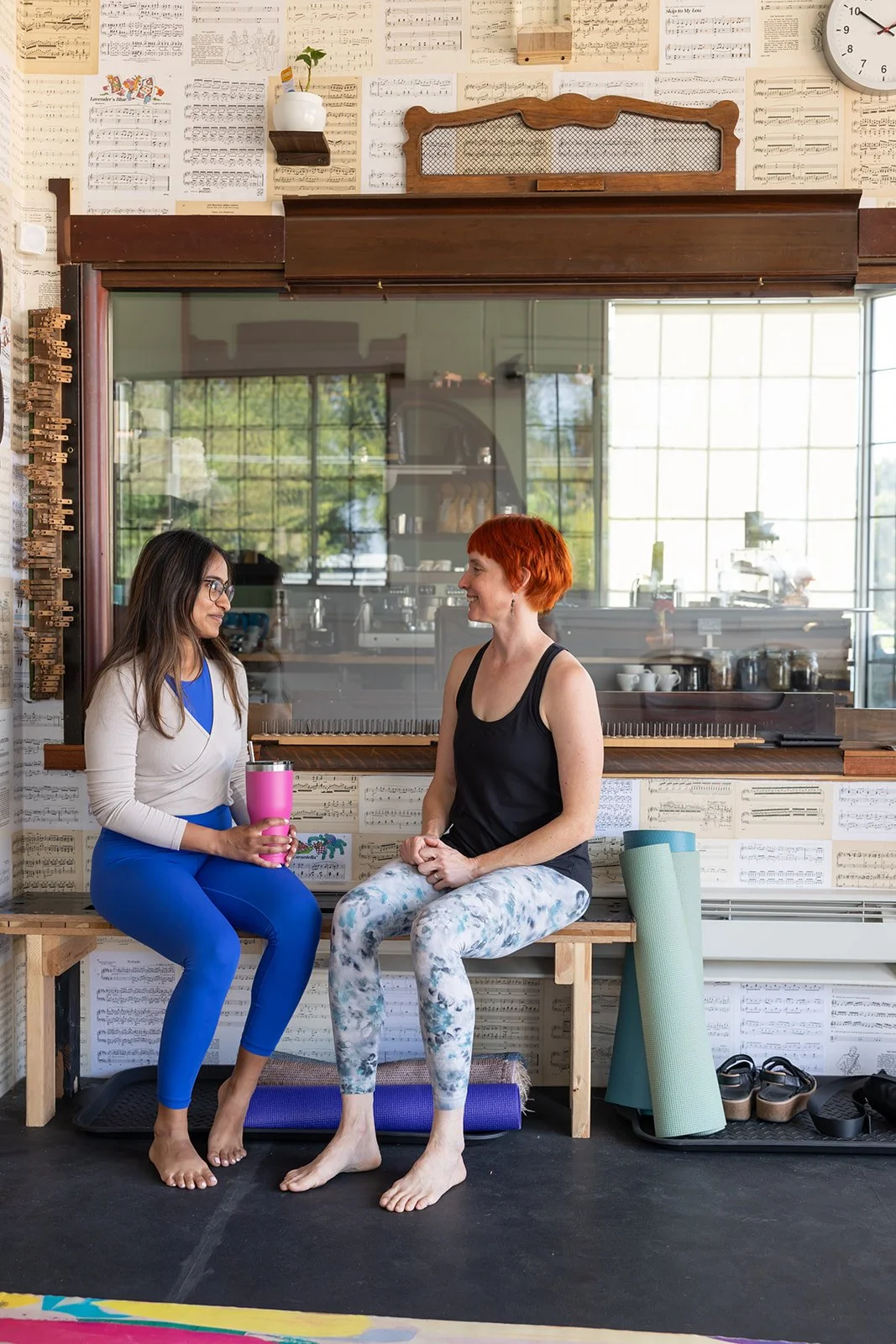 Two women sitting on a wooden bench in a yoga studio, smiling at each other, with yoga mats and shoes on the floor nearby, and music sheets on the wall behind them.