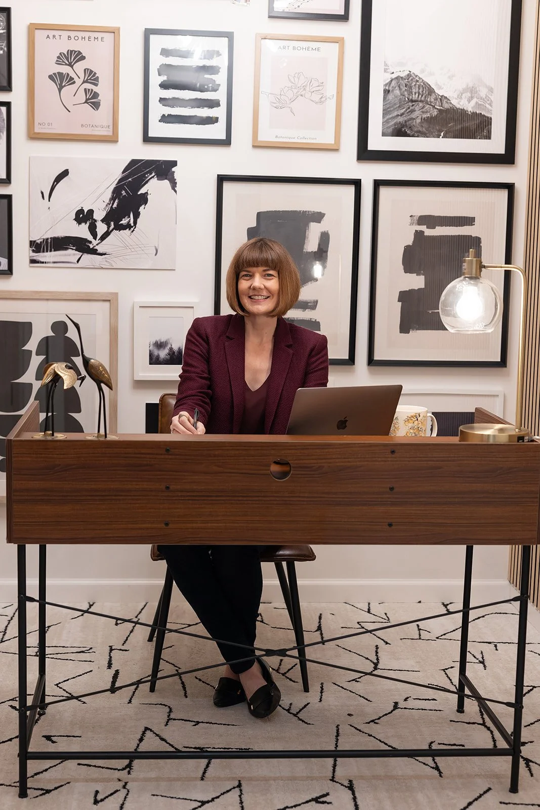 A woman sitting at a wooden desk with a laptop, smiling, in a room with a white wall decorated with black and white framed art.
