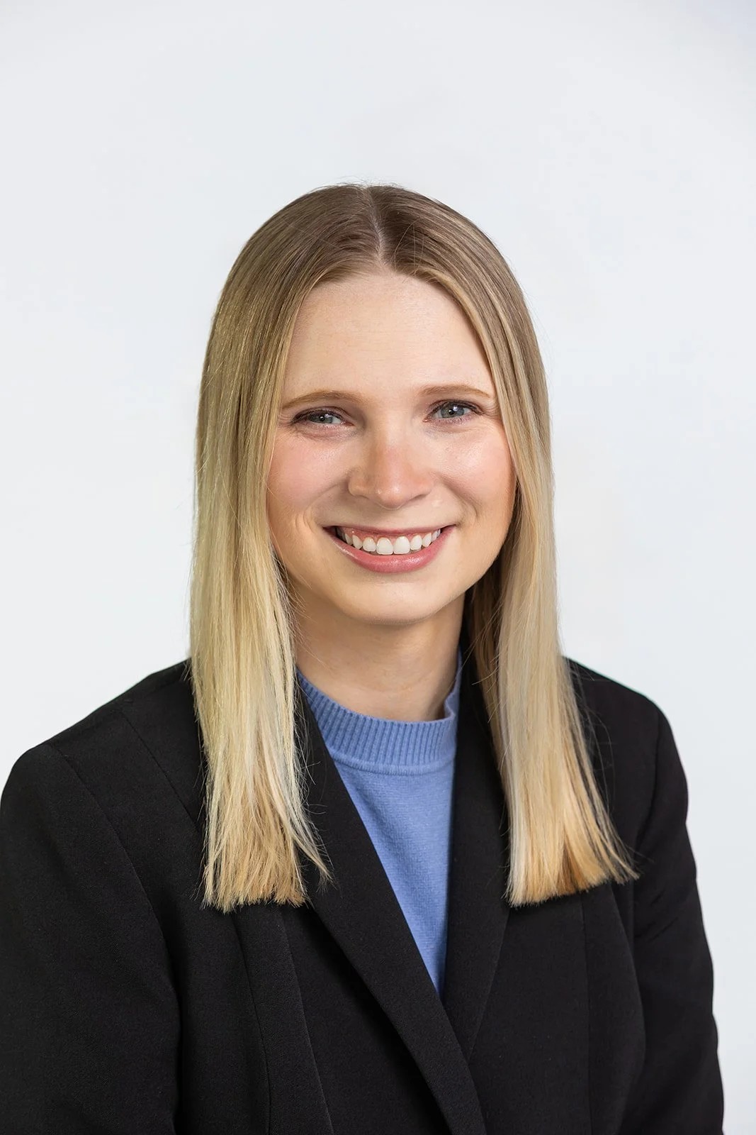 A young woman with blonde hair wearing a black blazer and a blue turtleneck, smiling against a plain white background.