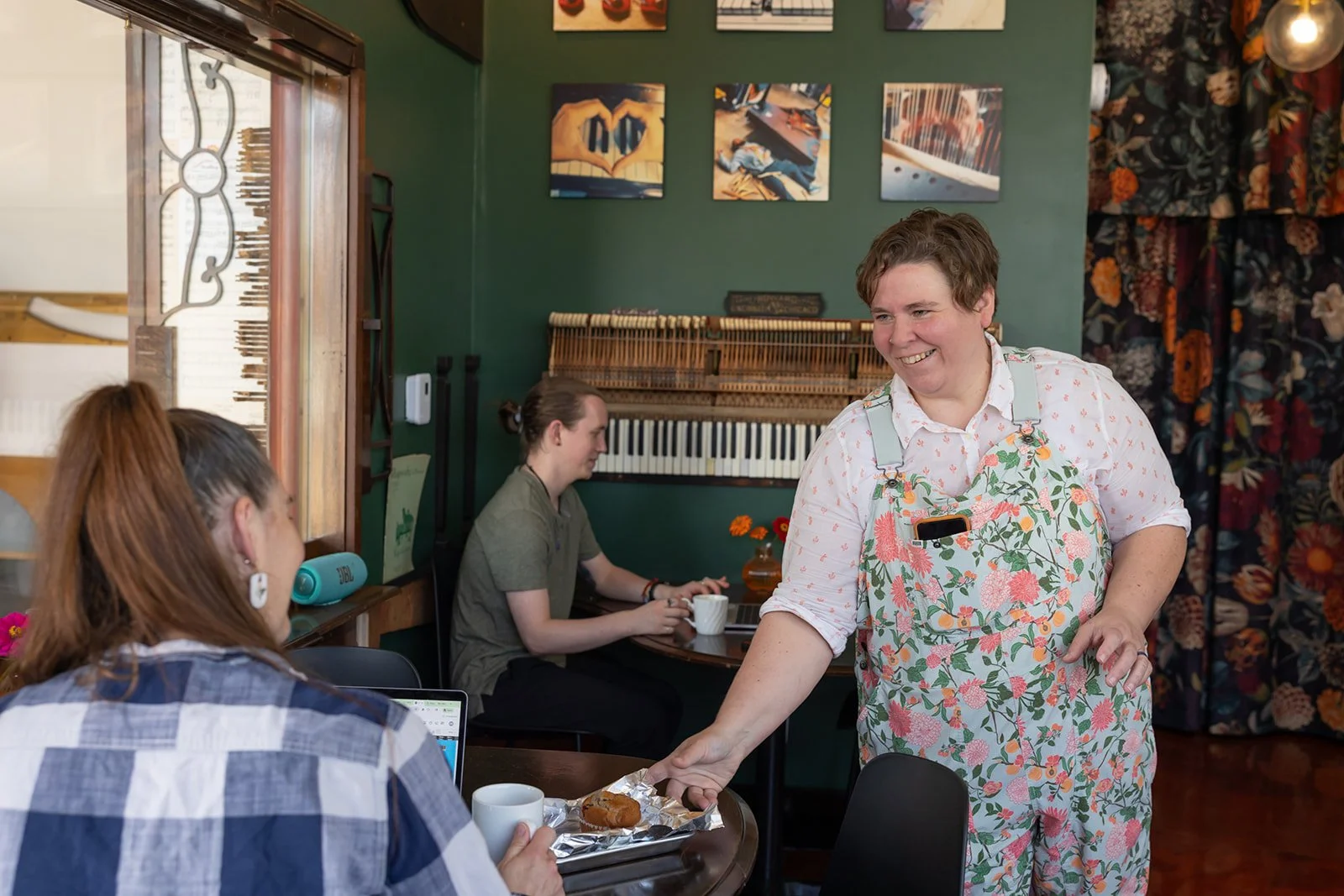 A woman with short hair wearing floral apron is serving a pastry and coffee to a seated customer inside a cozy cafe with dark green walls, musical instruments on the wall, and framed artwork.