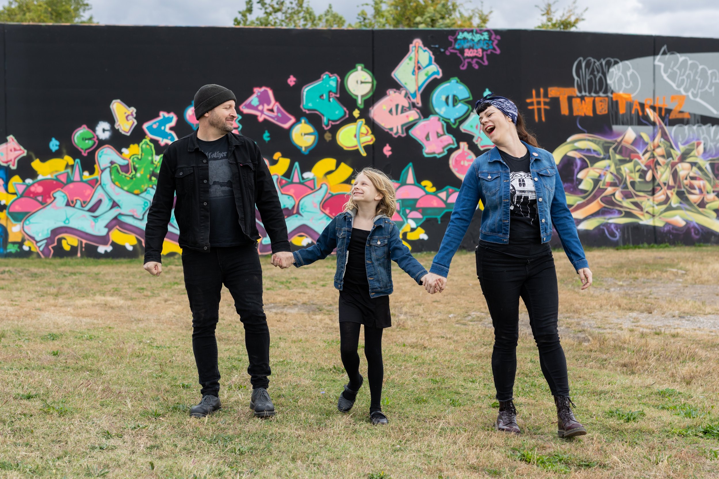A family of three holding hands and walking in a grassy area near a colorful graffiti wall. The man on the left is wearing a black beanie and black jacket, the girl in the middle is wearing a denim jacket and black dress, and the woman on the right i