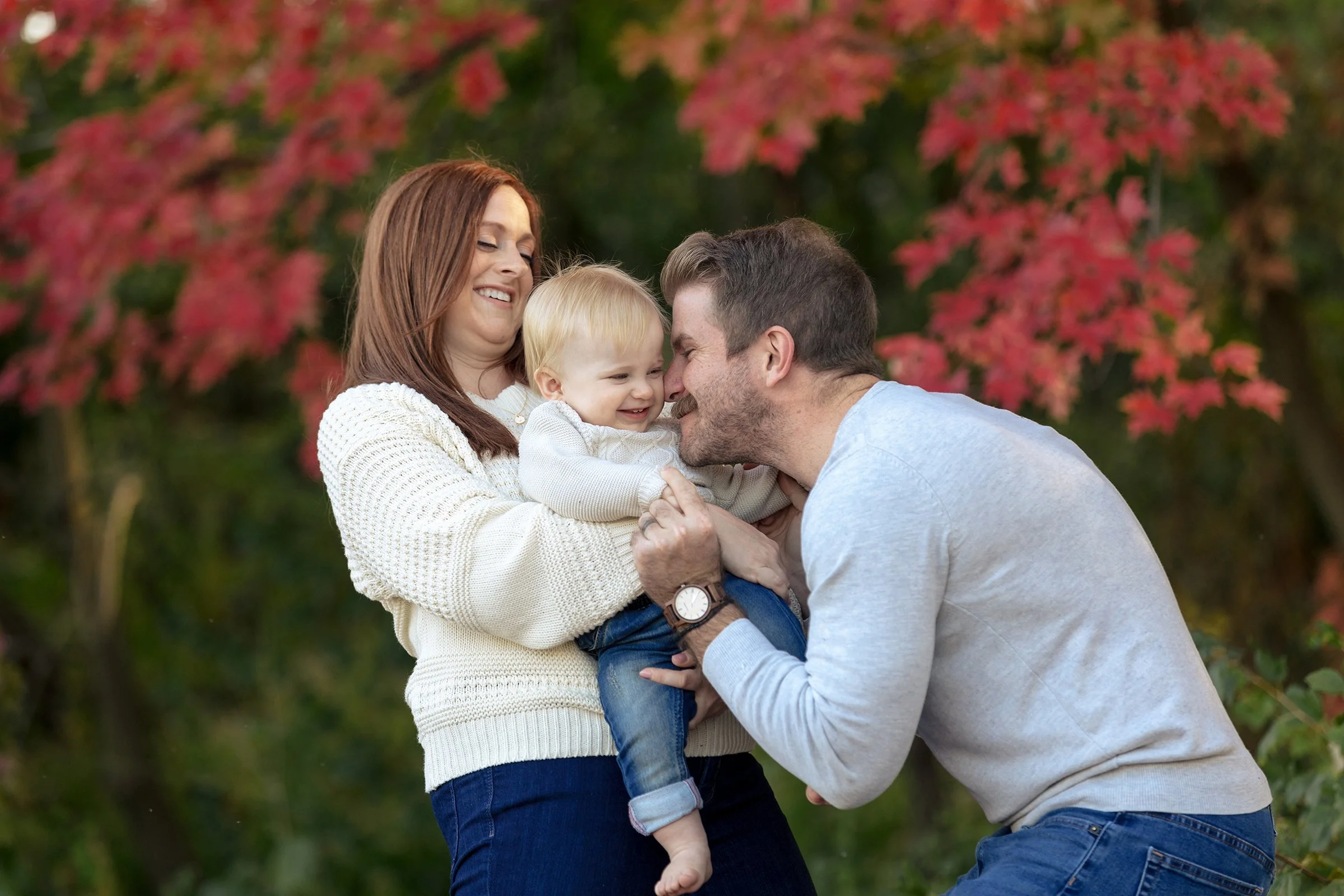 A family of three outdoors, with pink and green foliage in the background, smiling and playing together.
