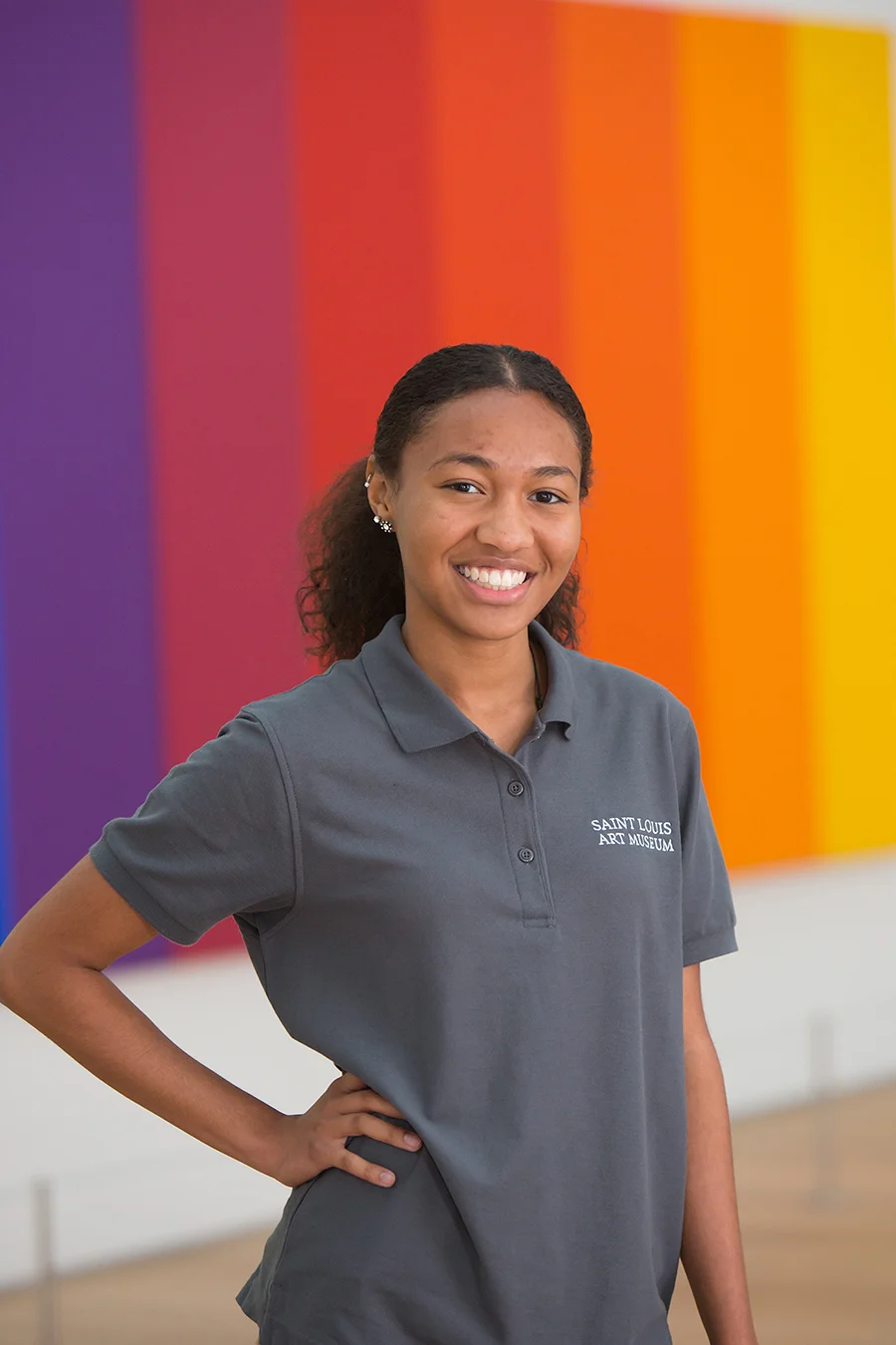 Smiling young woman wearing a gray polo shirt with 'Saint Louis Art Museum' logo, standing in front of a colorful, rainbow-like painted wall.