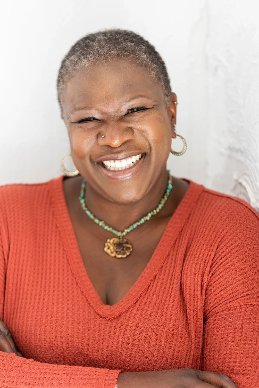 A smiling woman with short gray and black hair, wearing a rust-colored top, hoop earrings, a turquoise beaded necklace, and a wooden pendant necklace, standing against a white textured background.