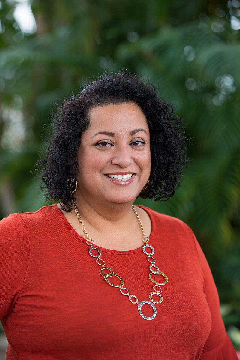 A woman with curly black hair, wearing a red top and a statement necklace, smiling outdoors with a blurred green background of trees or plants.